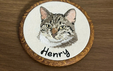 Custom embroidered pet hoop portrait of a tabby cat named Henry in natural wood hoop with metal hanger, featuring detailed gray and cream machine embroidery and personalized name lettering