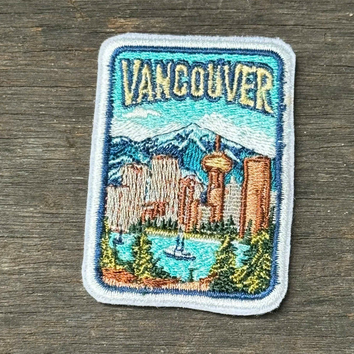 An embroidered rectangular patch featuring a colorful illustration of Vancouver, including the skyline, mountains, water, and the word 'Vancouver' at the top, placed on a dark wood surface.