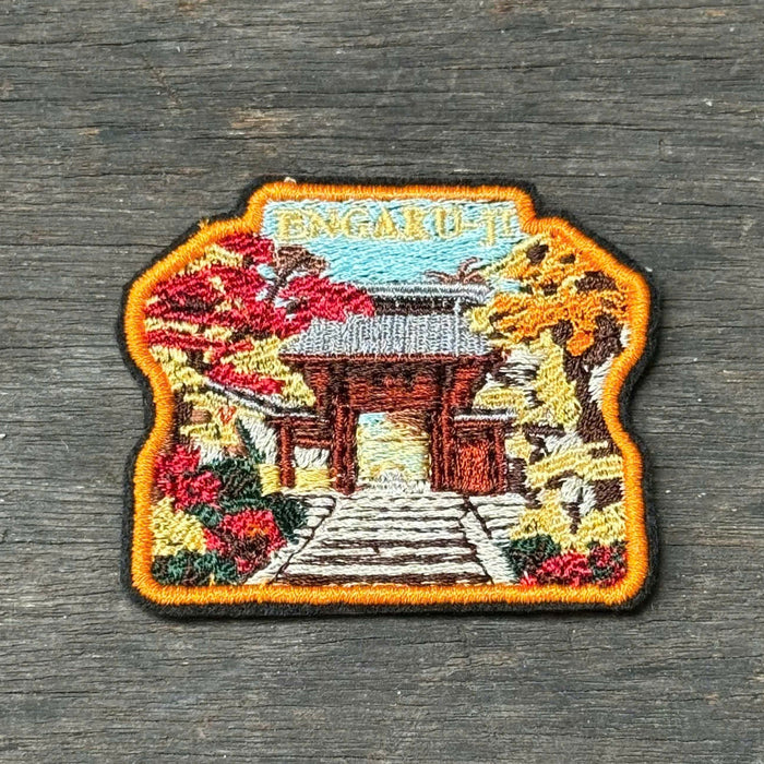 An embroidered patch depicting a Japanese temple gate surrounded by autumn foliage, placed on a dark gray wooden surface.
