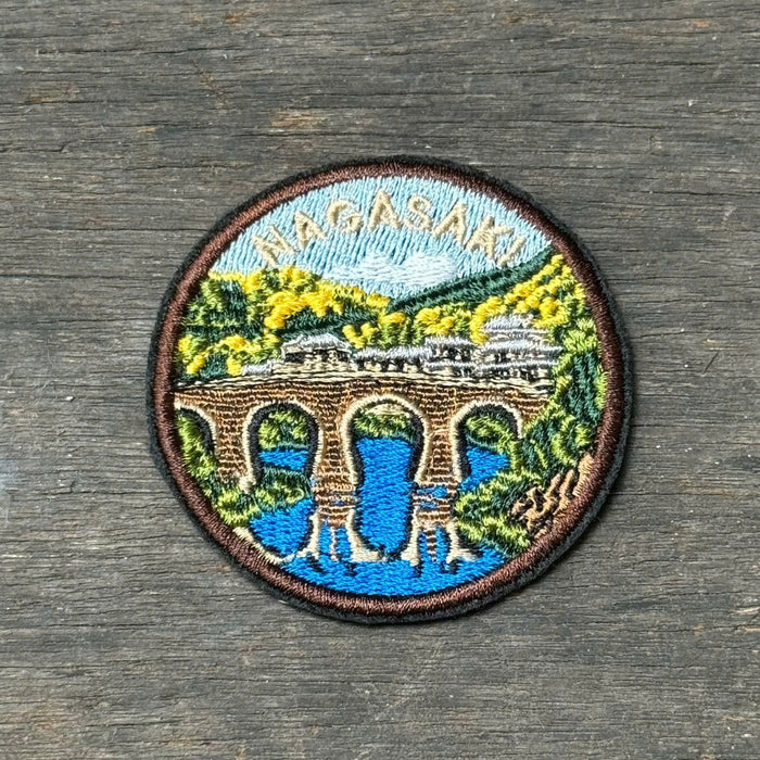 A circular embroidered patch depicting a scenic landscape with a bridge over blue water, green trees, and a light blue sky, placed on a dark gray wooden surface.