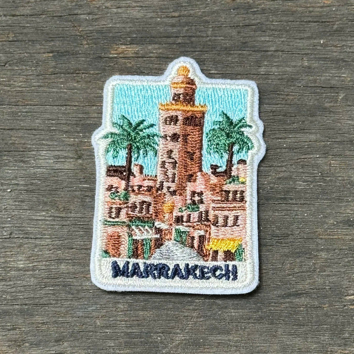 A small embroidered patch depicting a Moroccan Marrakech scene with a tower, palm trees, and the text “MARRAKECH,” placed on a dark wood surface.