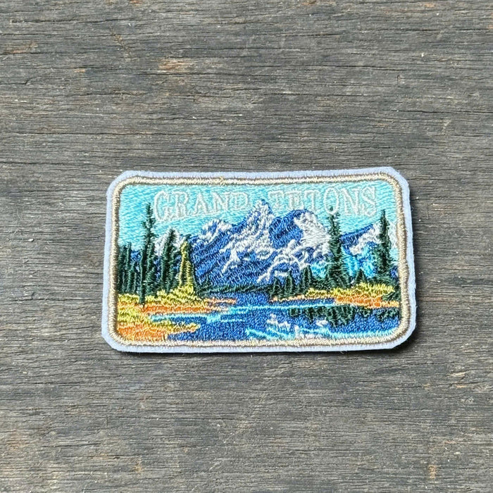 A rectangular embroidered patch depicting a mountain lake landscape with pine trees and the words 'Grand Teton', placed on a weathered gray wooden surface.