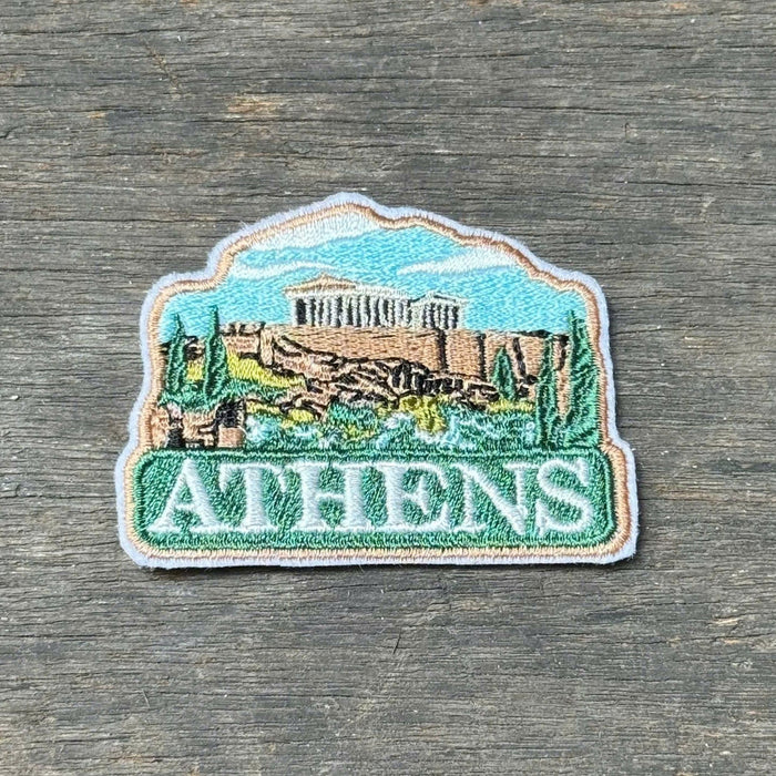 An embroidered patch featuring the Acropolis of Athens, with the word 'ATHENS' in white text on a green banner at the bottom, placed on a weathered gray wooden surface.