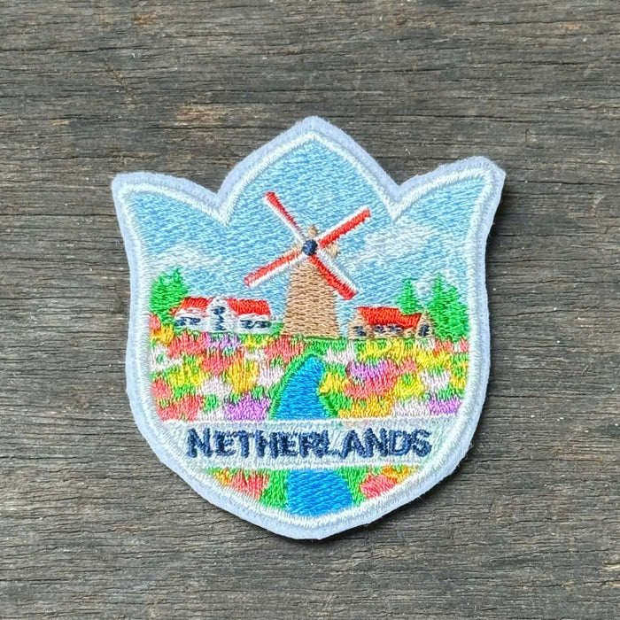 A small embroidered Netherlands patch shaped like a tulip, featuring a windmill, flowers, and the word 'NETHERLANDS,' placed on a dark weathered wood surface.