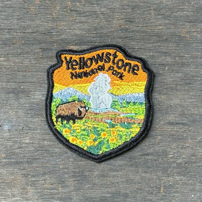 An embroidered shield-shaped patch featuring Yellowstone National Park scenery with a bison, steamboat, mountains, and yellow flowers, placed on a weathered gray wooden surface.