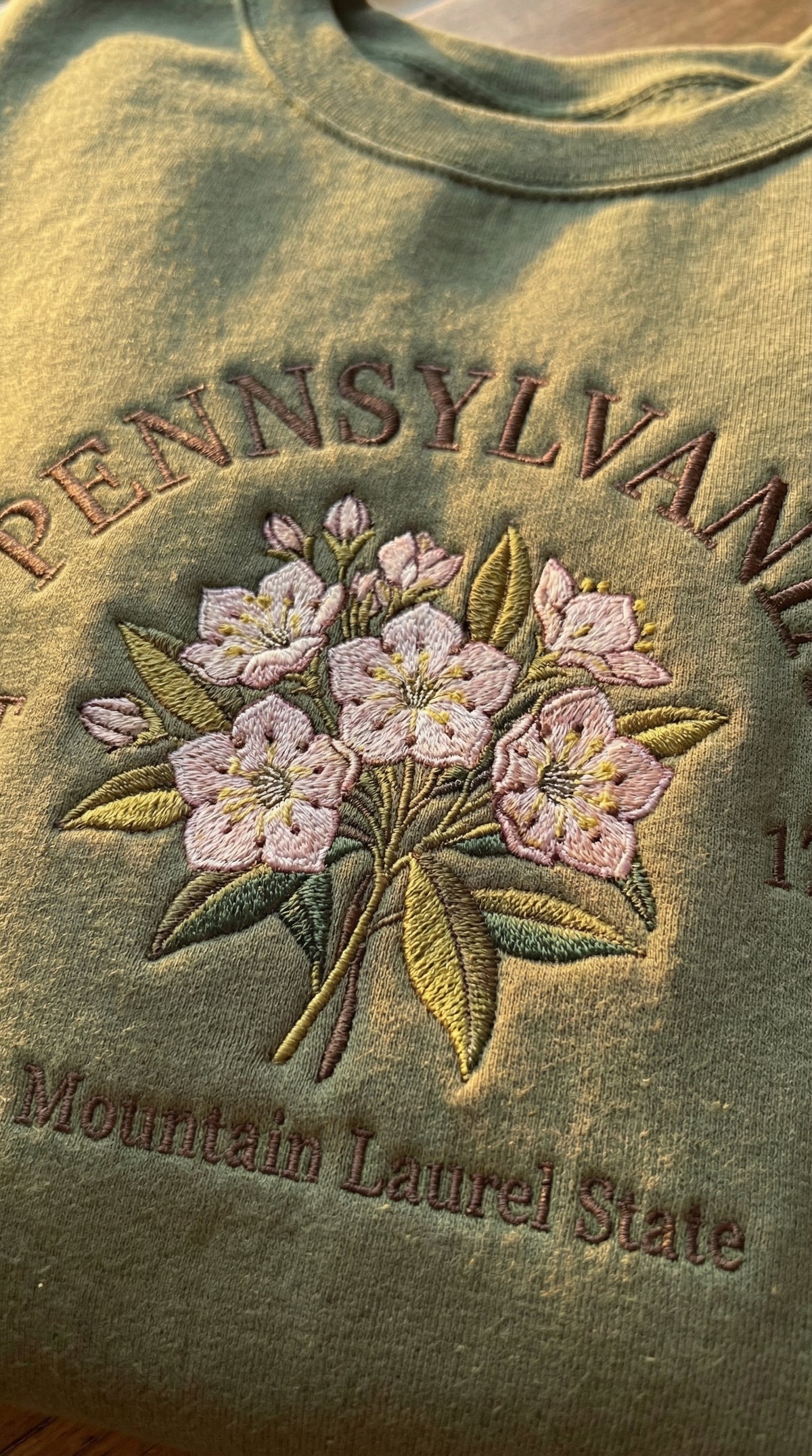 Olive green folded sweatshirt with detailed pink Mountain Laurel flower embroidery reading Pennsylvania EST 1787 The Mountain Laurel State