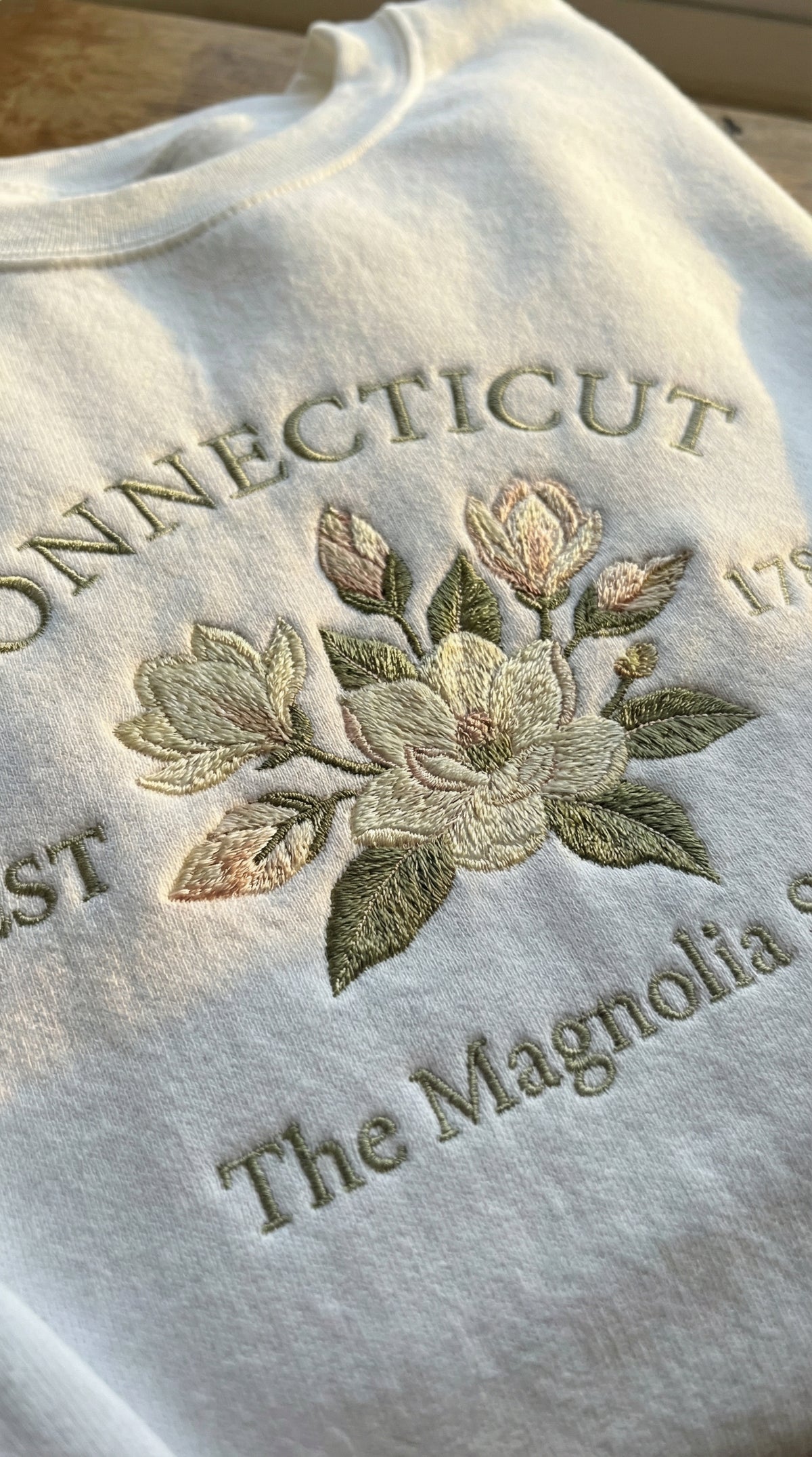 White folded unisex sweatshirt featuring detailed pastel magnolia flower embroidery, with text reading Connecticut EST 1788 The Magnolia State