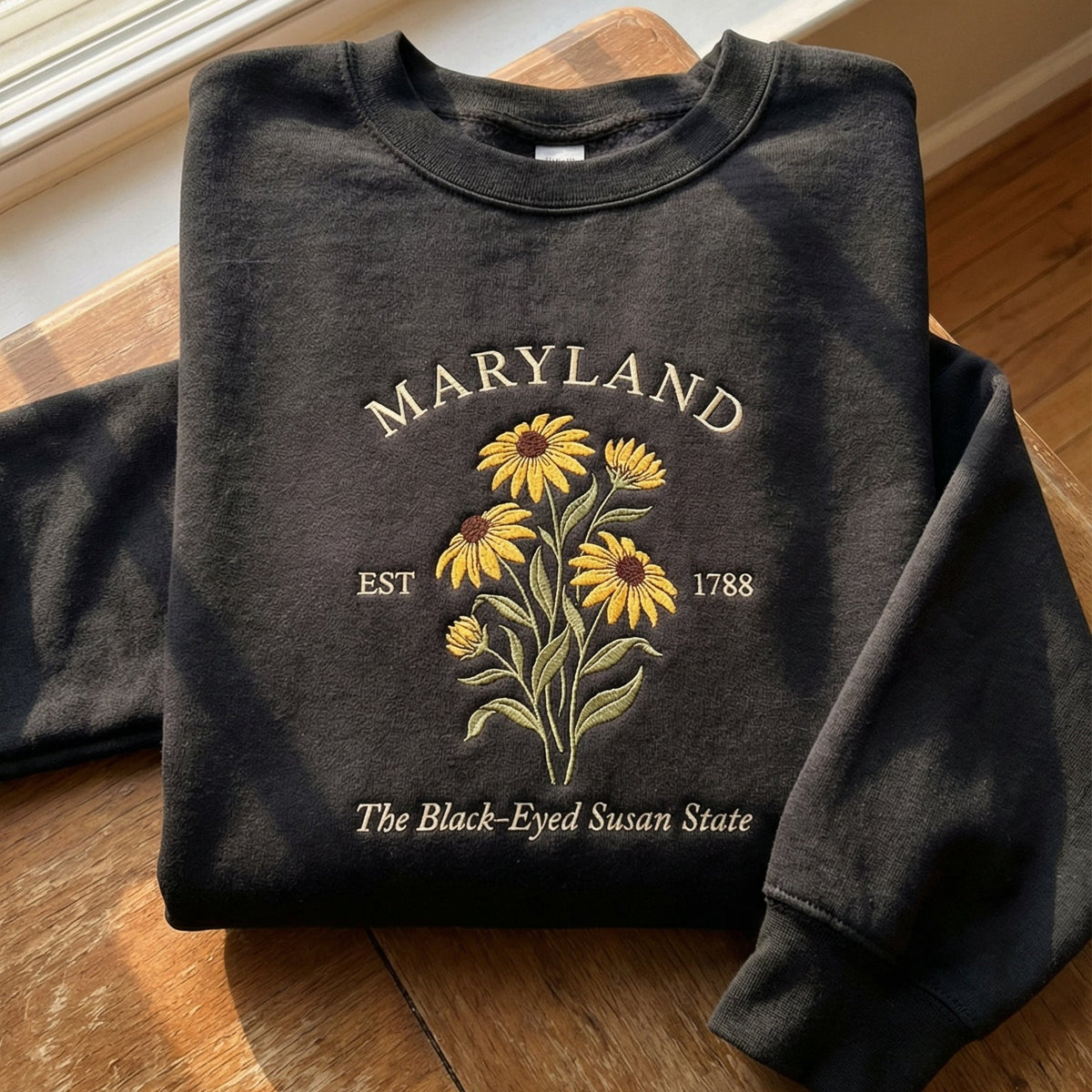 Black folded sweatshirt with detailed yellow Black-Eyed Susan flower embroidery reading Maryland EST 1788 The Black-Eyed Susan State