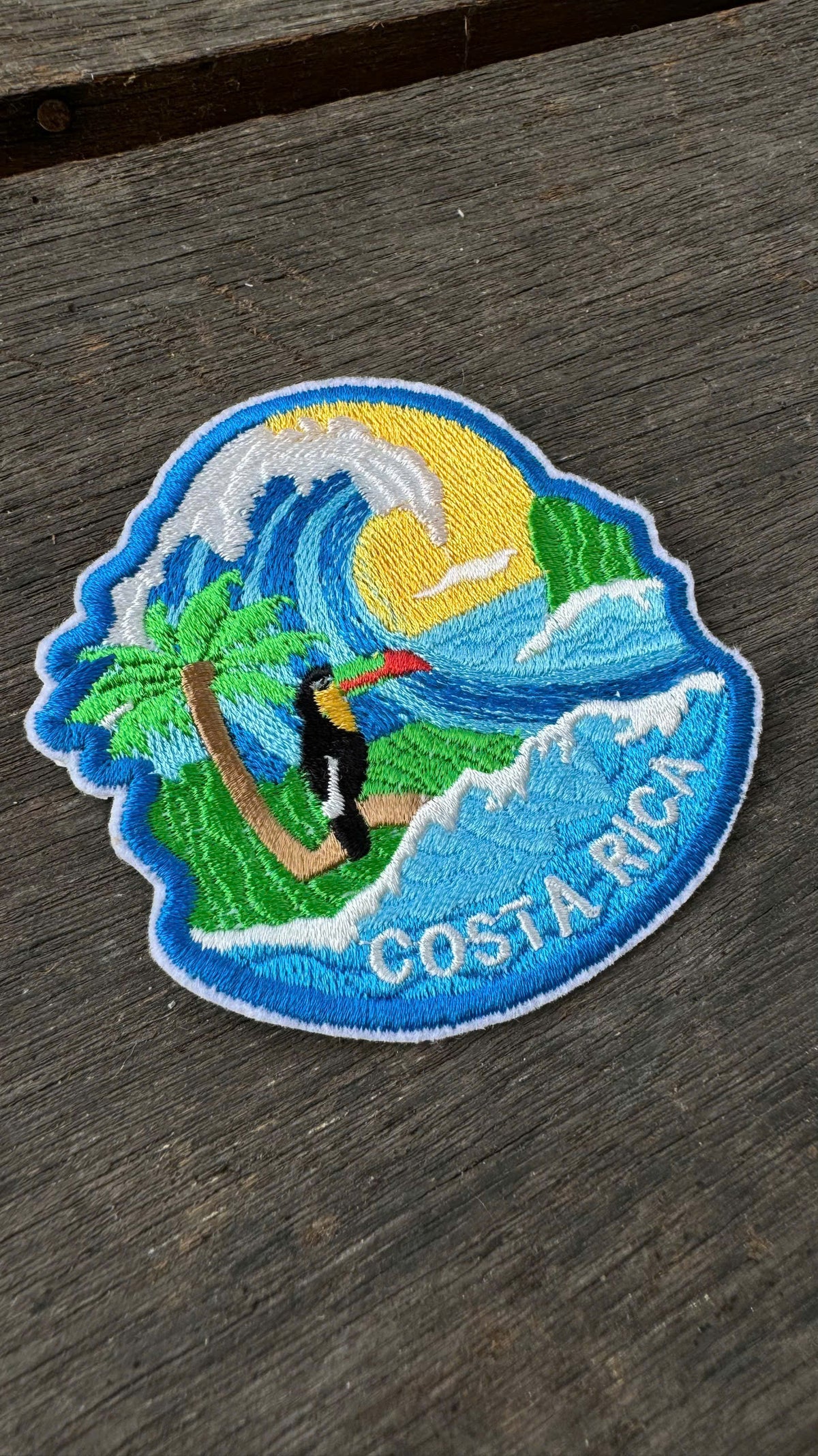 Embroidered patch of Costa Rica with a toucan, wave, and palm tree on a wooden surface.