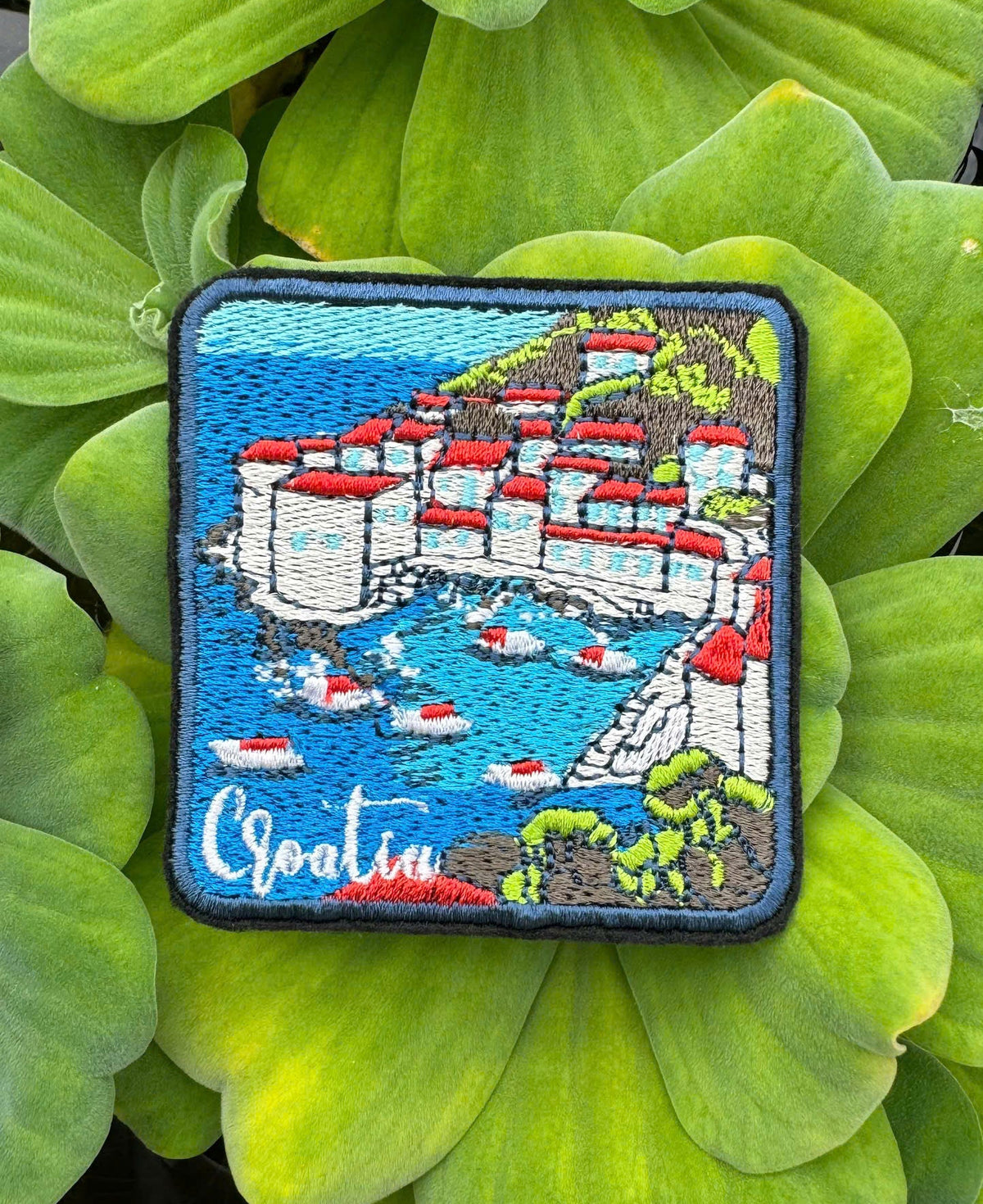 Embroidered patch of a coastal scene with buildings and water, placed on green leaves.