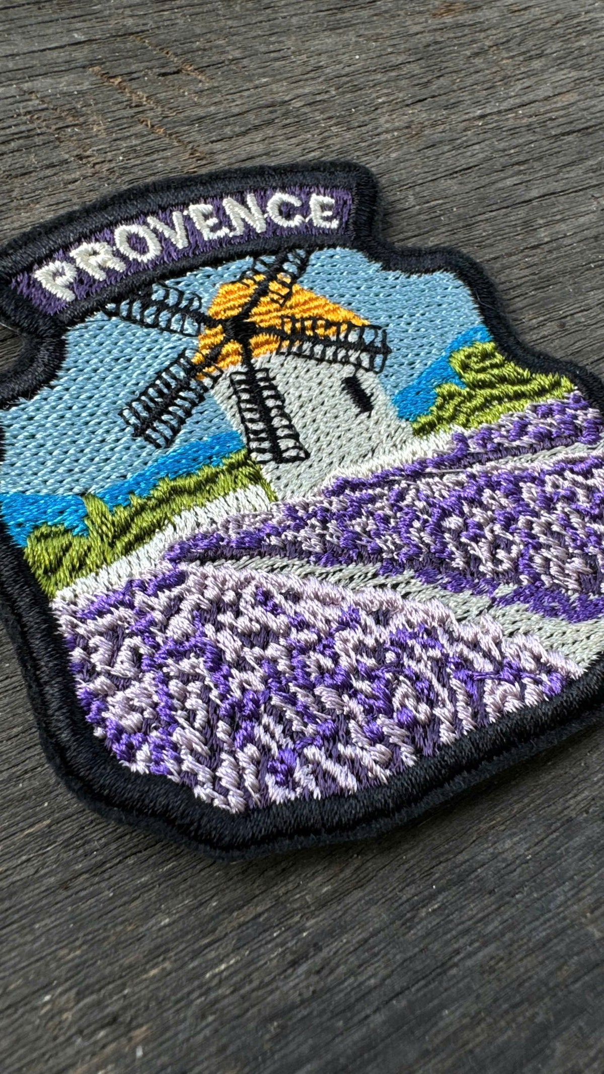 Embroidered patch with a windmill and lavender field labeled 'Provence' on a wooden surface.