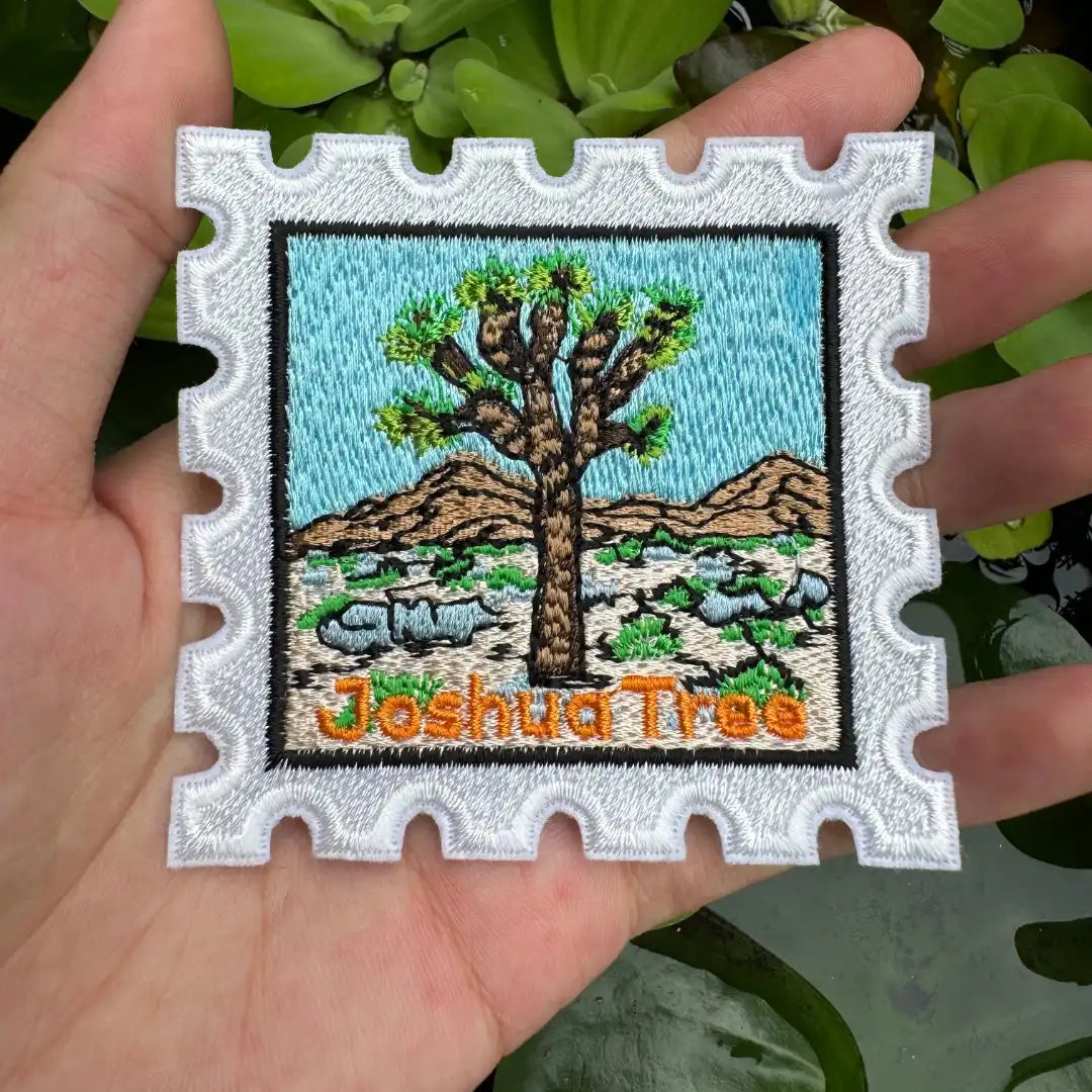 Embroidered patch of a Joshua Tree with 'Joshua Tree' text held in a hand against a green leafy background.