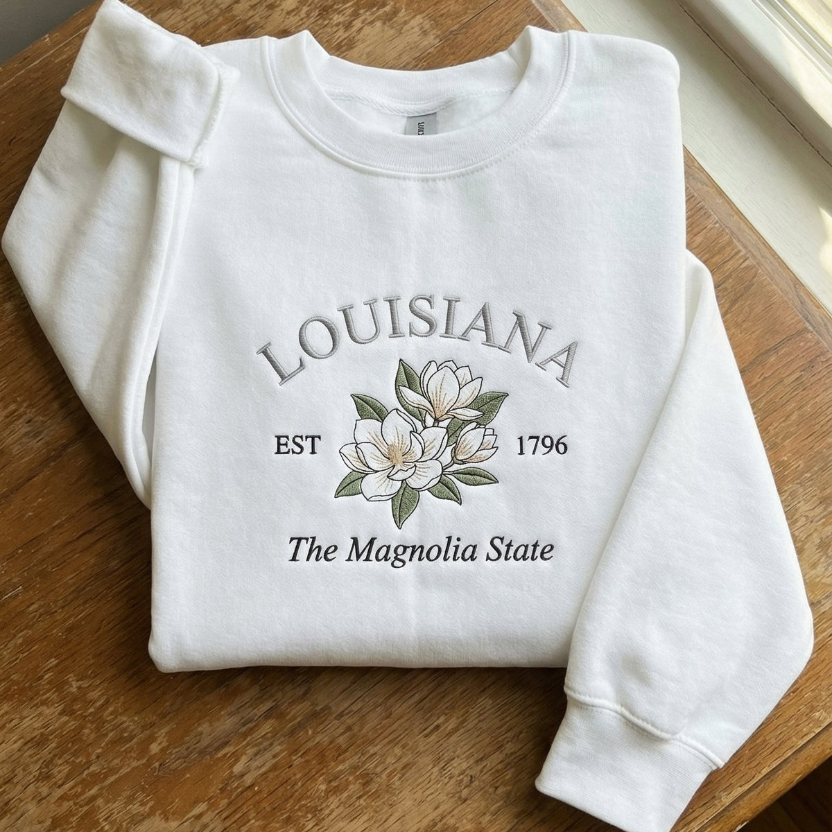 White folded sweatshirt with detailed Magnolia flower embroidery reading Louisiana EST 1796 The Magnolia State