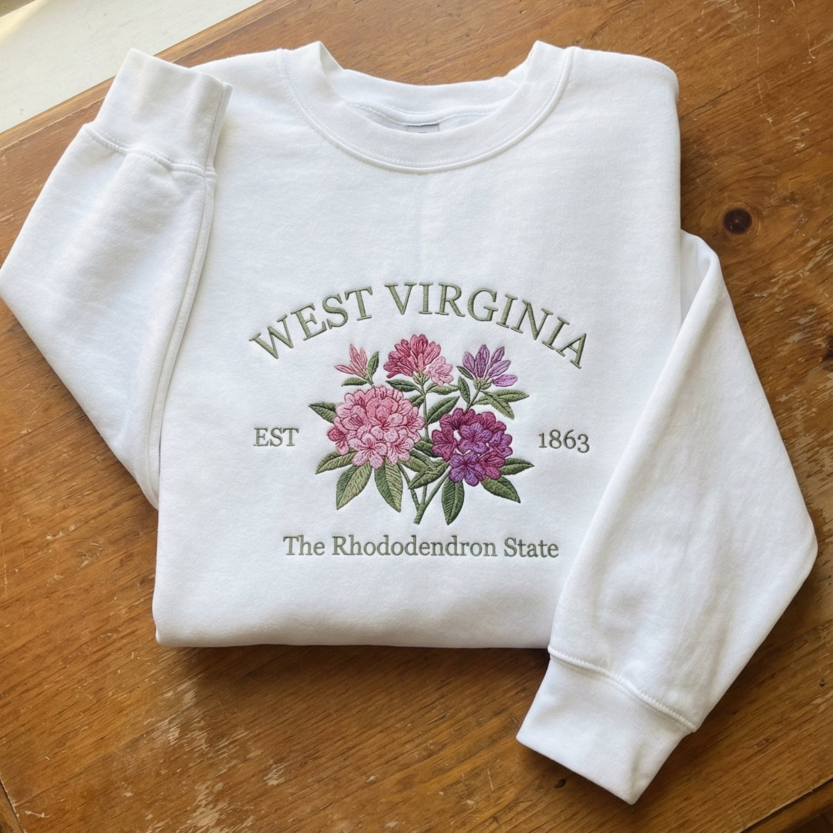White folded sweatshirt with detailed pink and purple Rhododendron flower embroidery, reading West Virginia EST 1863 The Rhododendron State