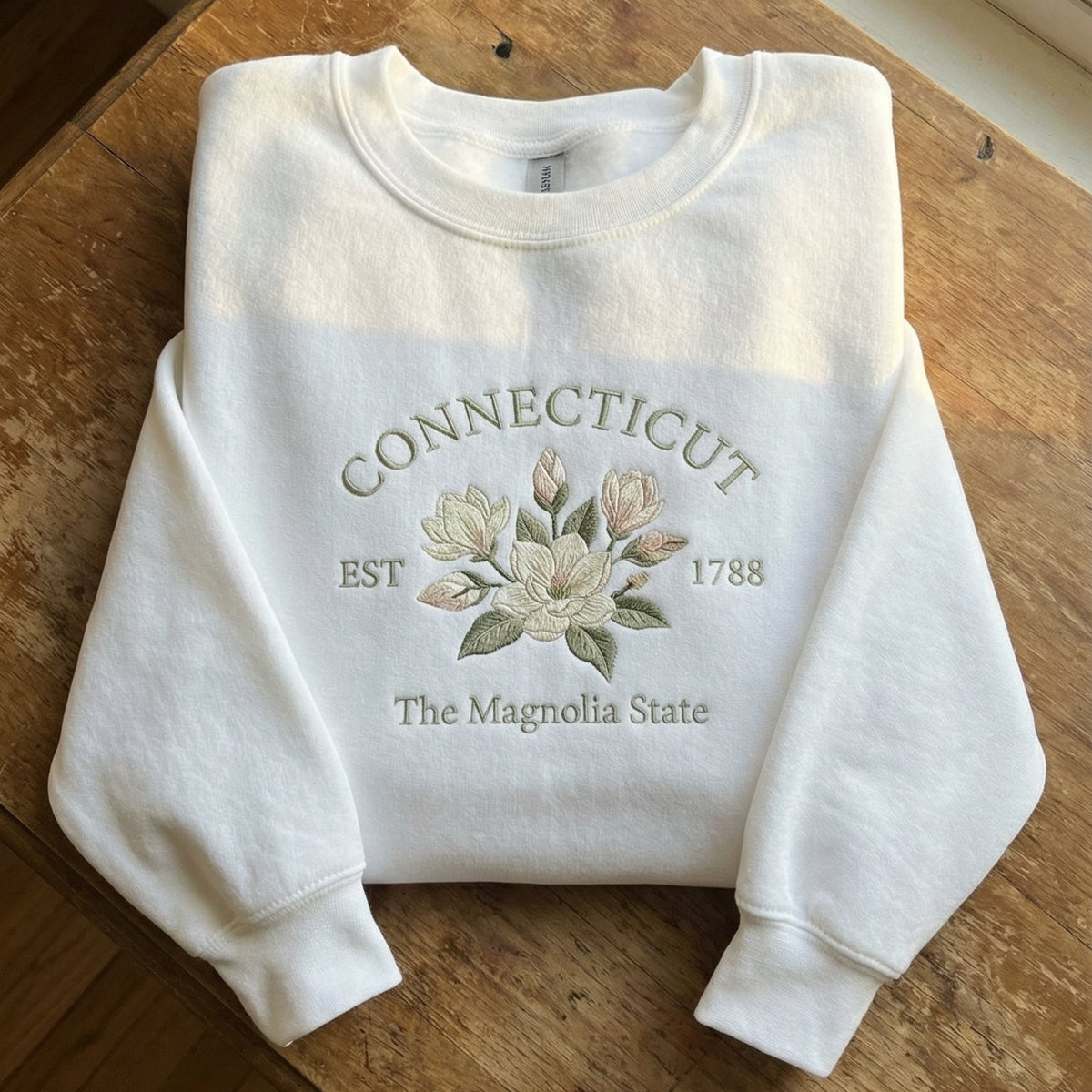White folded unisex sweatshirt featuring detailed pastel magnolia flower embroidery, with text reading Connecticut EST 1788 The Magnolia State