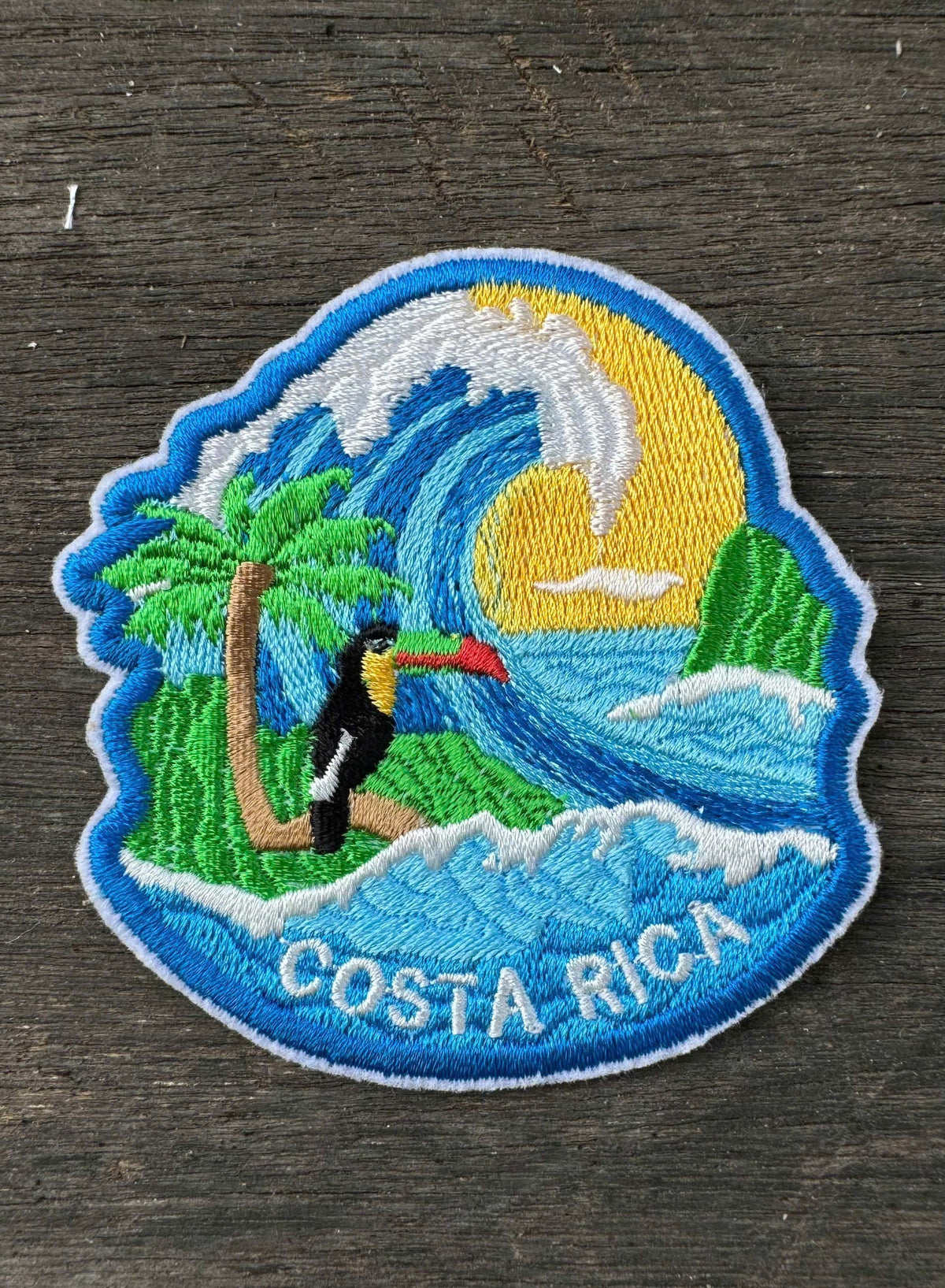 Embroidered patch of Costa Rica with a toucan, palm tree, and wave on a wooden surface.