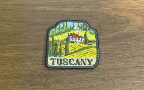 Machine-embroidered Tuscany travel patch showing yellow sunflower fields, red villa roof, green cypress trees, blue sky, and 'TUSCANY' text on cream background with gray border.
