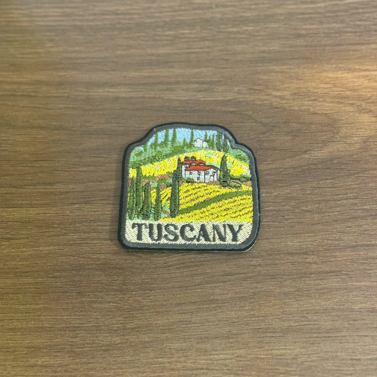 Machine-embroidered Tuscany travel patch showing yellow sunflower fields, red villa roof, green cypress trees, blue sky, and 'TUSCANY' text on cream background with gray border.