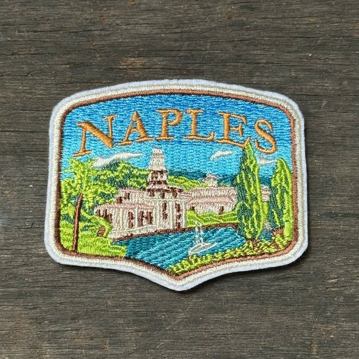 An embroidered 'NAPLES' patch featuring a scenic illustration of a historic building, trees, and blue water, placed on a dark wood surface.