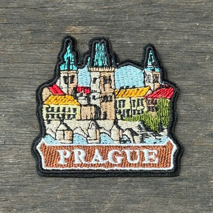 An embroidered patch depicting Prague's skyline with the word 'PRAGUE' on a banner, placed on a dark wood background.