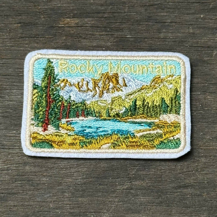 A rectangular embroidered patch depicting a mountain lake landscape with trees, placed on a dark wood surface.