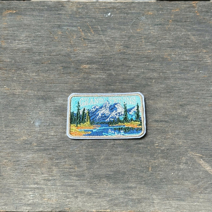 A rectangular embroidered patch depicting a mountain lake landscape with pine trees and the words 'Grand Teton', placed on a weathered gray wooden surface.