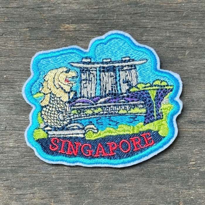 An embroidered Singapore patch featuring the Merlion and Marina Bay Sands, with 'SINGAPORE' stitched in red at the bottom, placed on a dark wood surface.