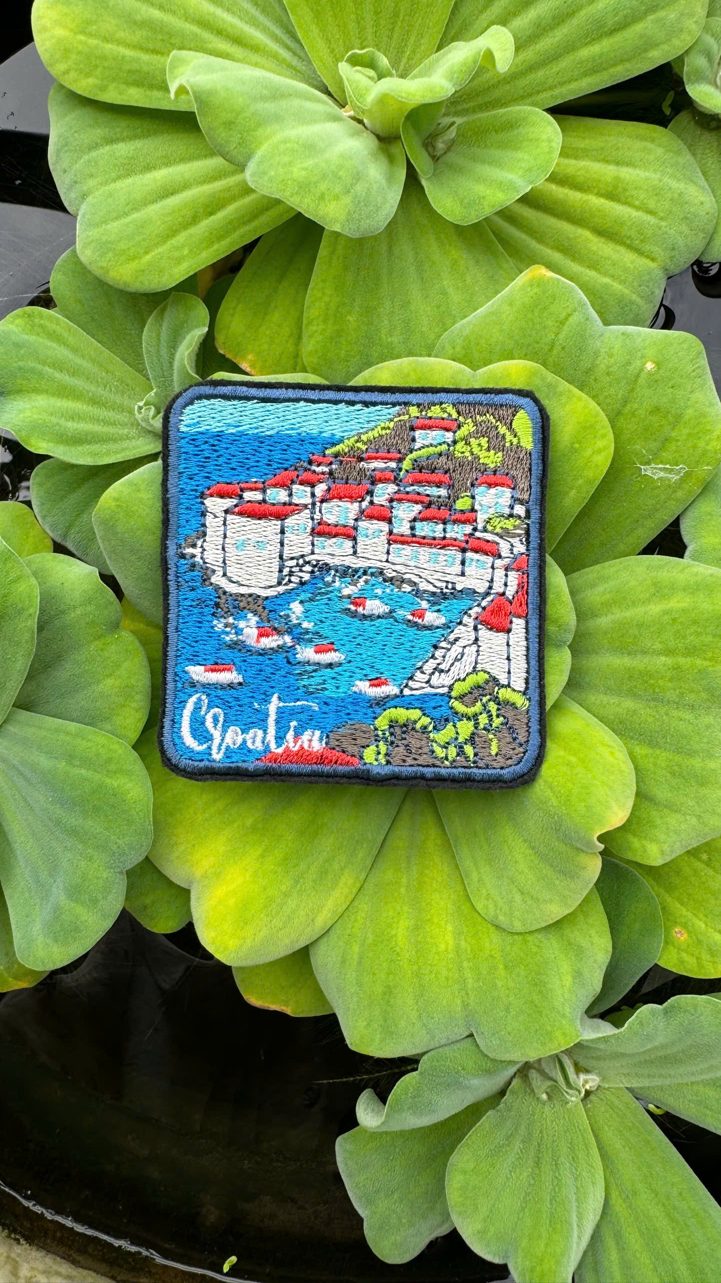 Embroidered patch of a coastal scene with buildings and water, placed on green leaves.