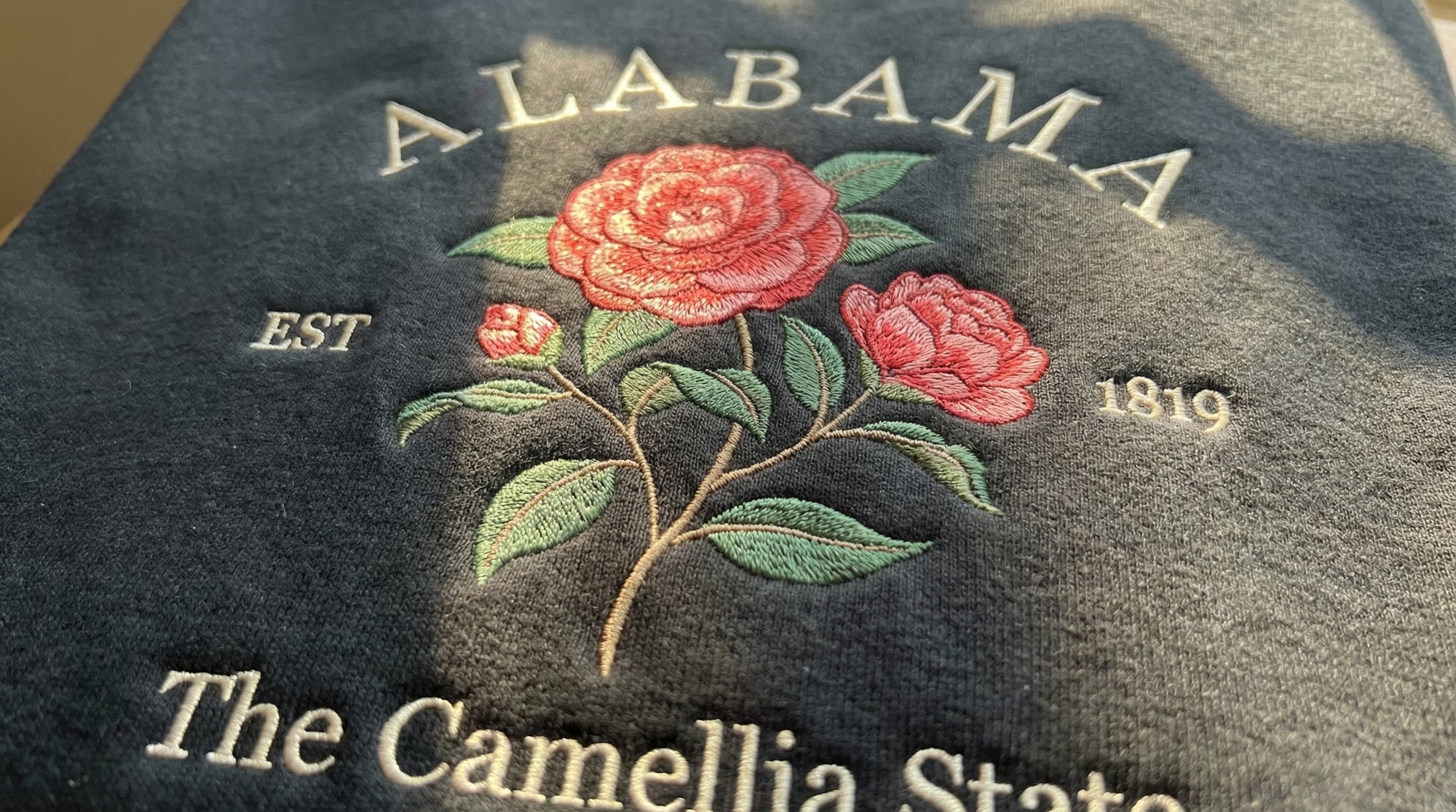 Black folded sweatshirt with detailed pink Camellia flower embroidery reading Alabama EST 1819 The Camellia State