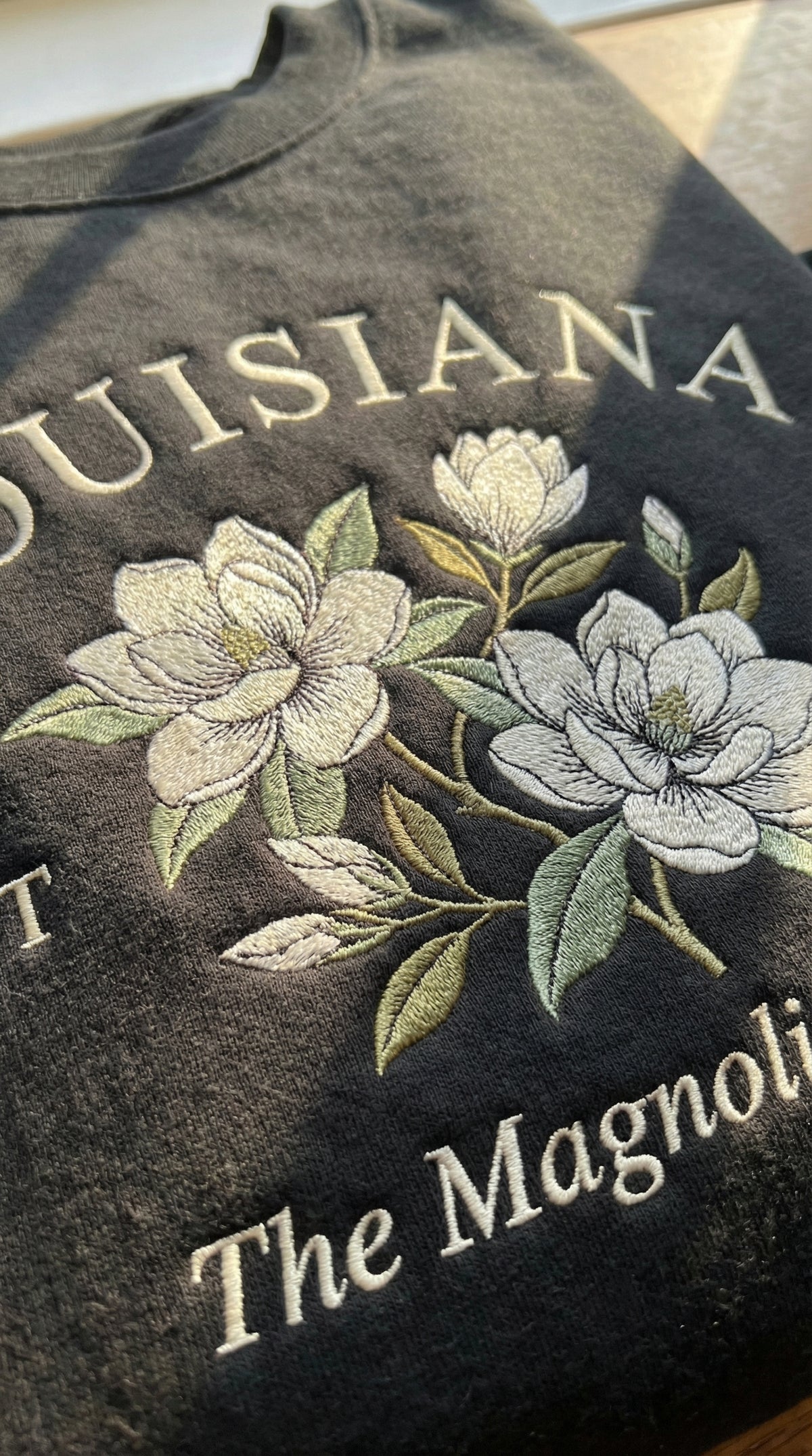 Black folded sweatshirt with detailed white Magnolia flower embroidery reading Louisiana EST 1796 The Magnolia State