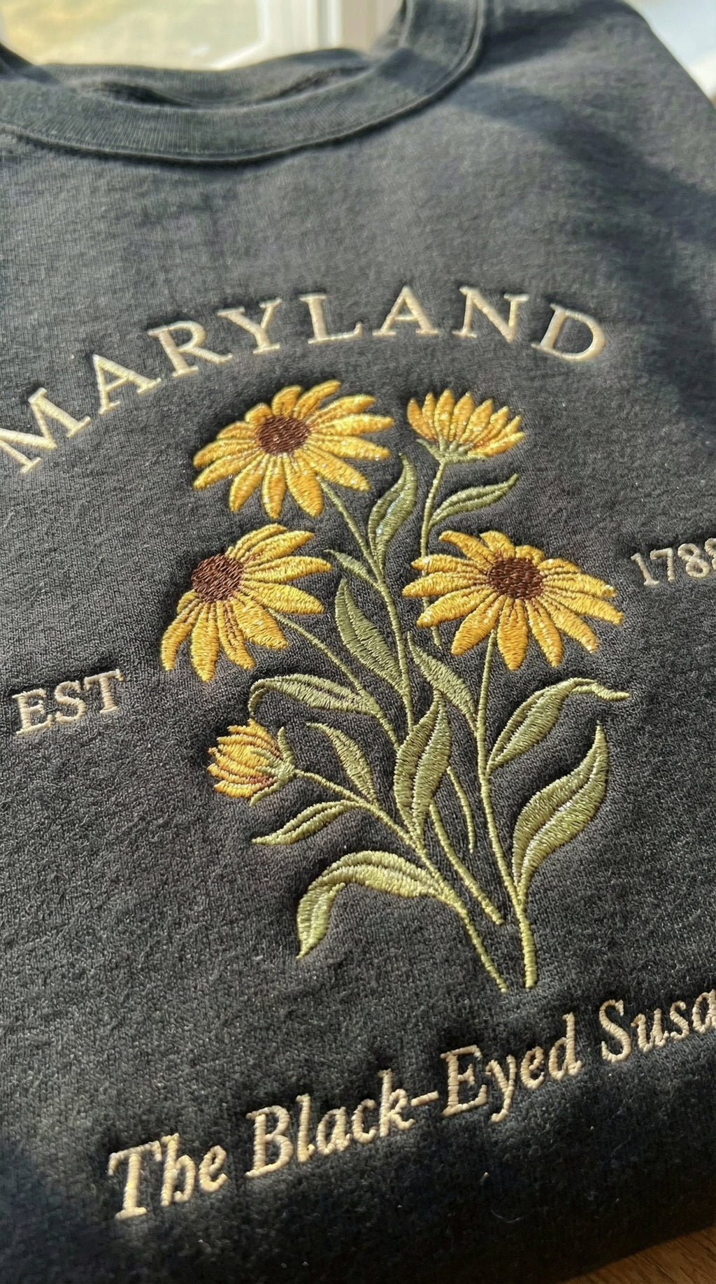 Black folded sweatshirt with detailed yellow Black-Eyed Susan flower embroidery reading Maryland EST 1788 The Black-Eyed Susan State