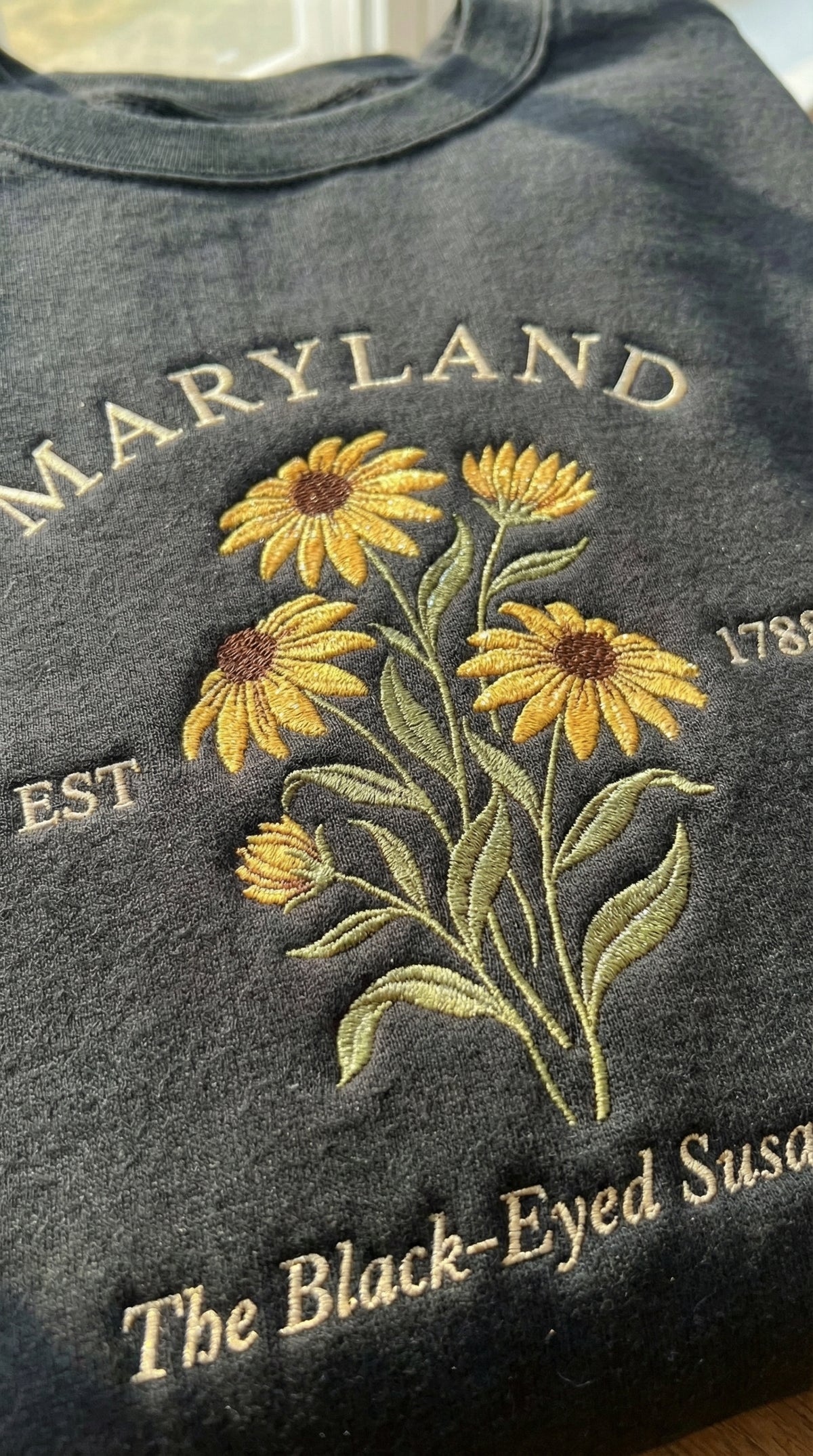 Black folded sweatshirt with detailed yellow Black-Eyed Susan flower embroidery reading Maryland EST 1788 The Black-Eyed Susan State