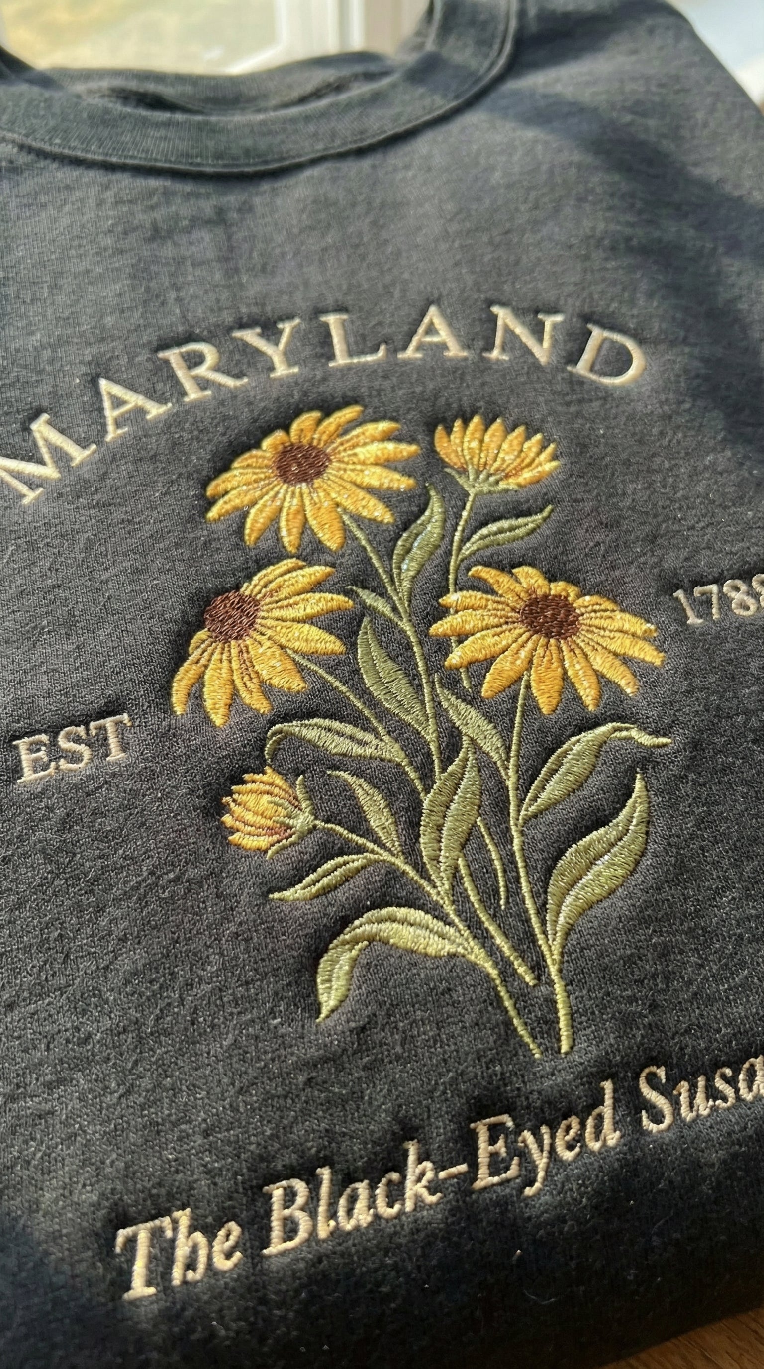 Black folded sweatshirt with detailed yellow Black-Eyed Susan flower embroidery reading Maryland EST 1788 The Black-Eyed Susan State