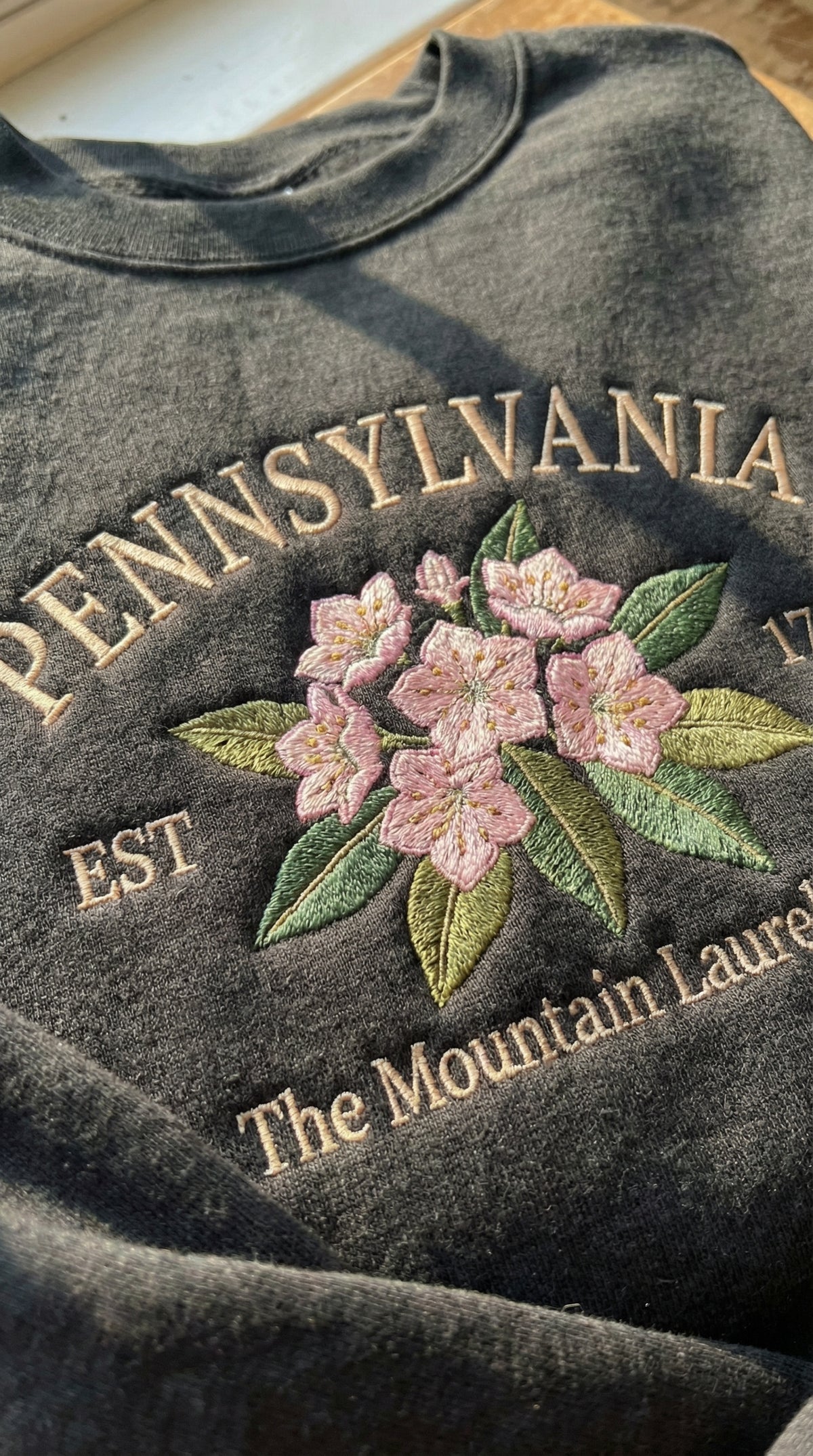 Charcoal black sweatshirt with detailed pink Mountain Laurel flower embroidery, reading Pennsylvania EST 1787 The Mountain Laurel State