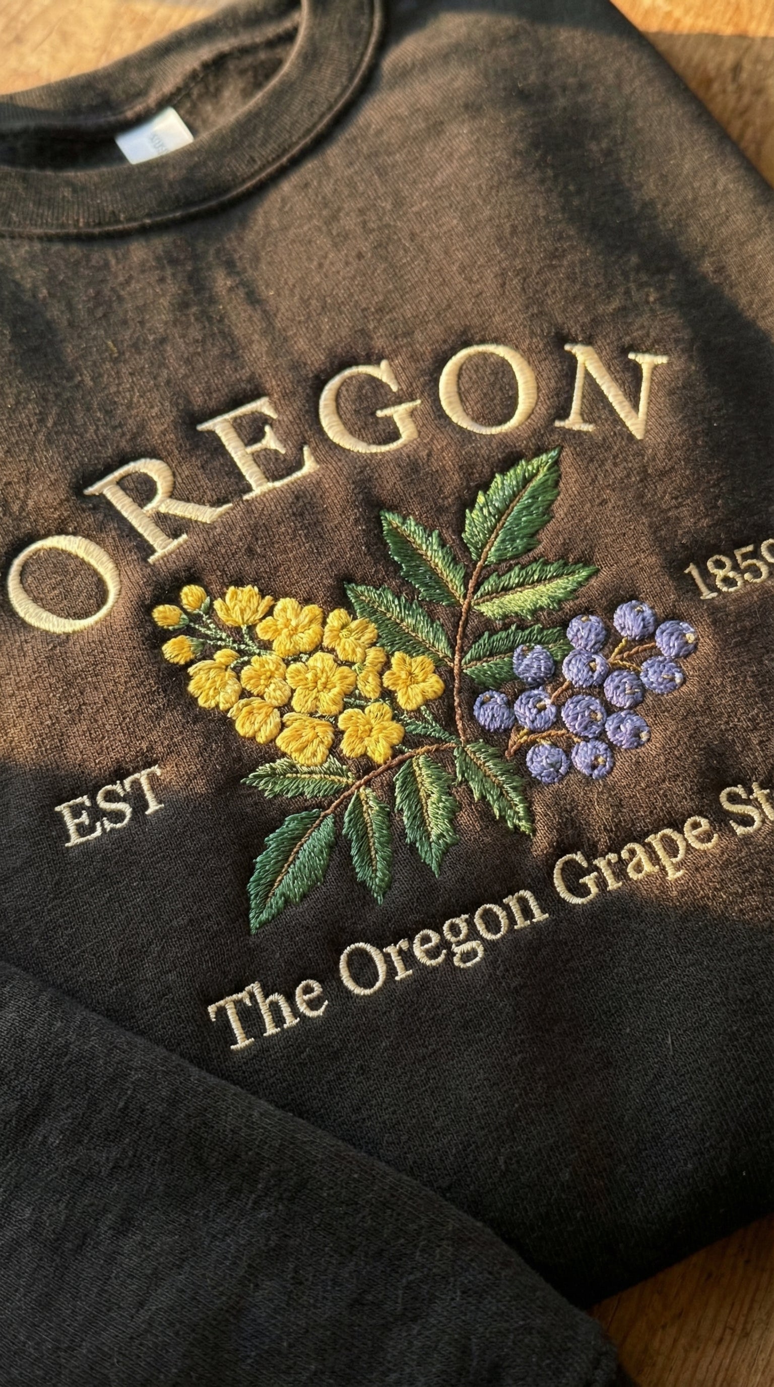 Charcoal black sweatshirt with detailed yellow Oregon Grape flowers and purple berries embroidery, reading Oregon EST 1859 The Oregon Grape State