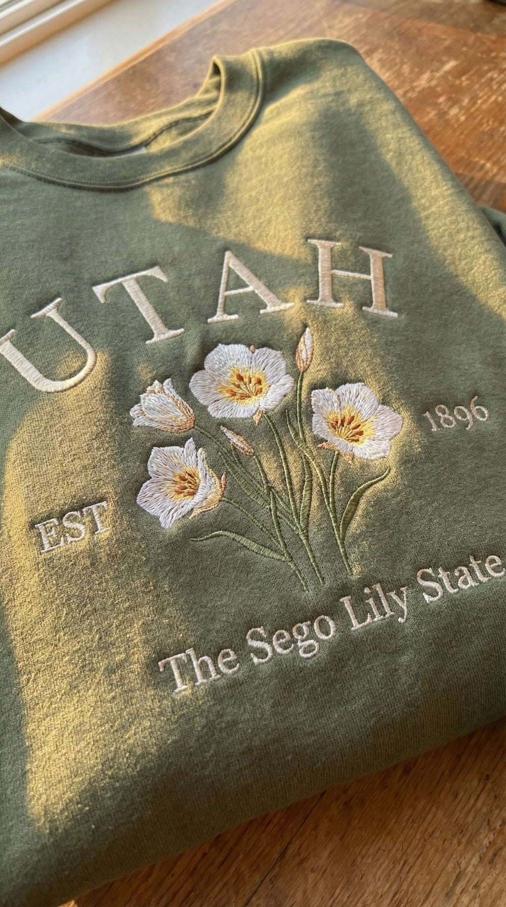 Olive green folded sweatshirt with detailed white Sego Lily flower embroidery reading Utah EST 1896 The Sego Lily State