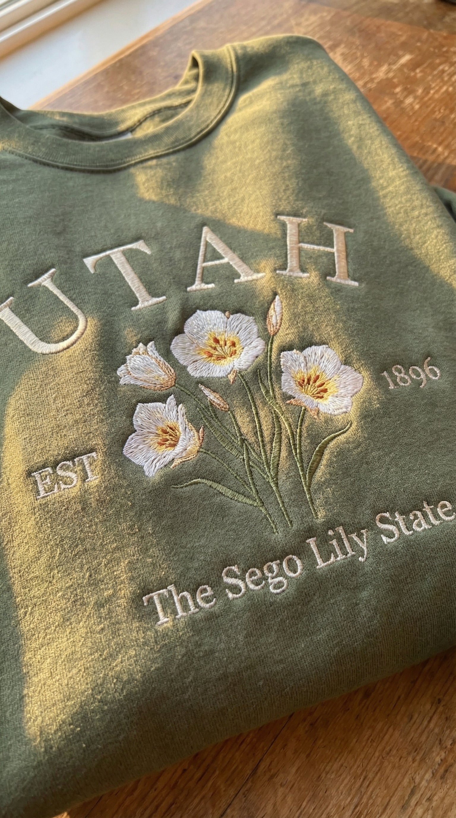 Olive green folded sweatshirt with detailed white Sego Lily flower embroidery reading Utah EST 1896 The Sego Lily State