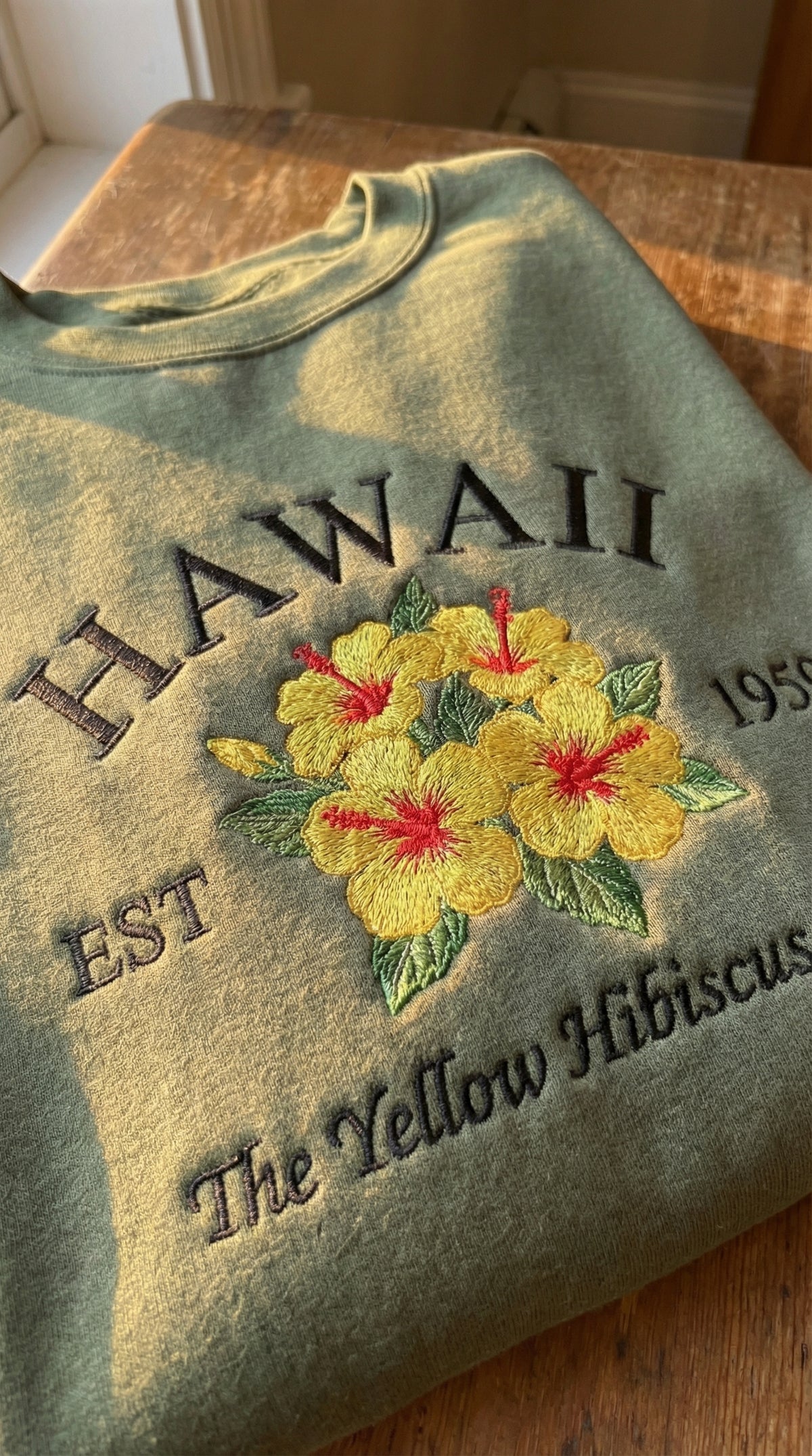 Olive green folded sweatshirt with detailed yellow hibiscus flower and text embroidery 'Hawaii EST 1959 The Yellow Hibiscus State'