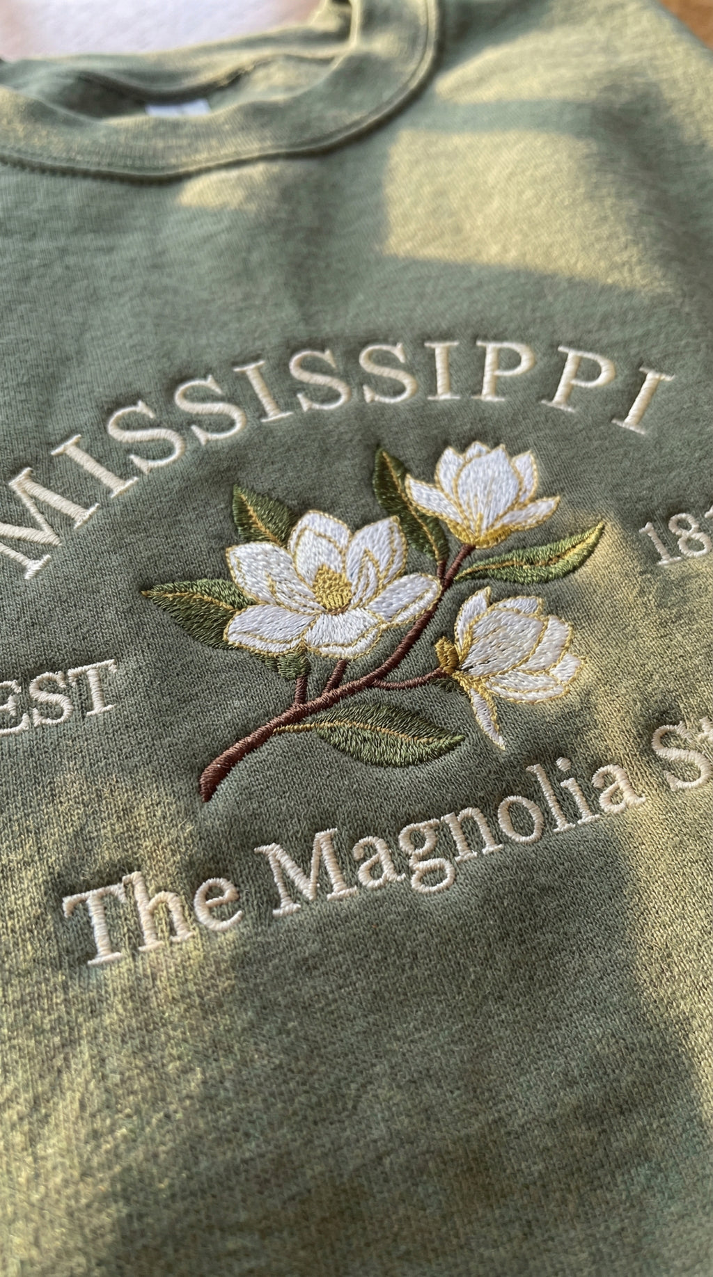 Olive green sweatshirt with detailed white Magnolia flower embroidery reading Mississippi EST 1817 The Magnolia State