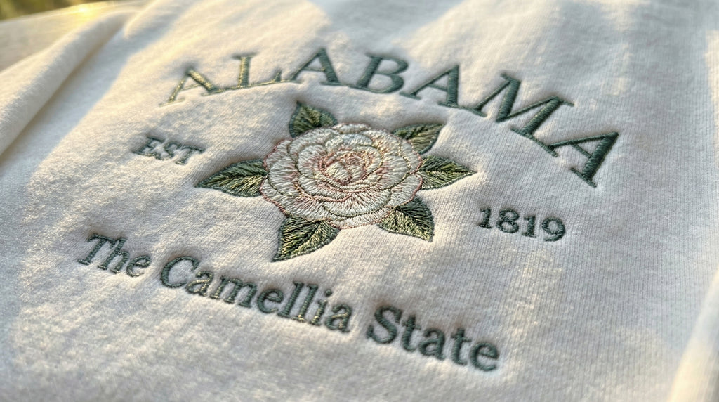 White folded sweatshirt with detailed pink Camellia flower embroidery and a pink banner reading Alabama EST 1819 The Camellia State