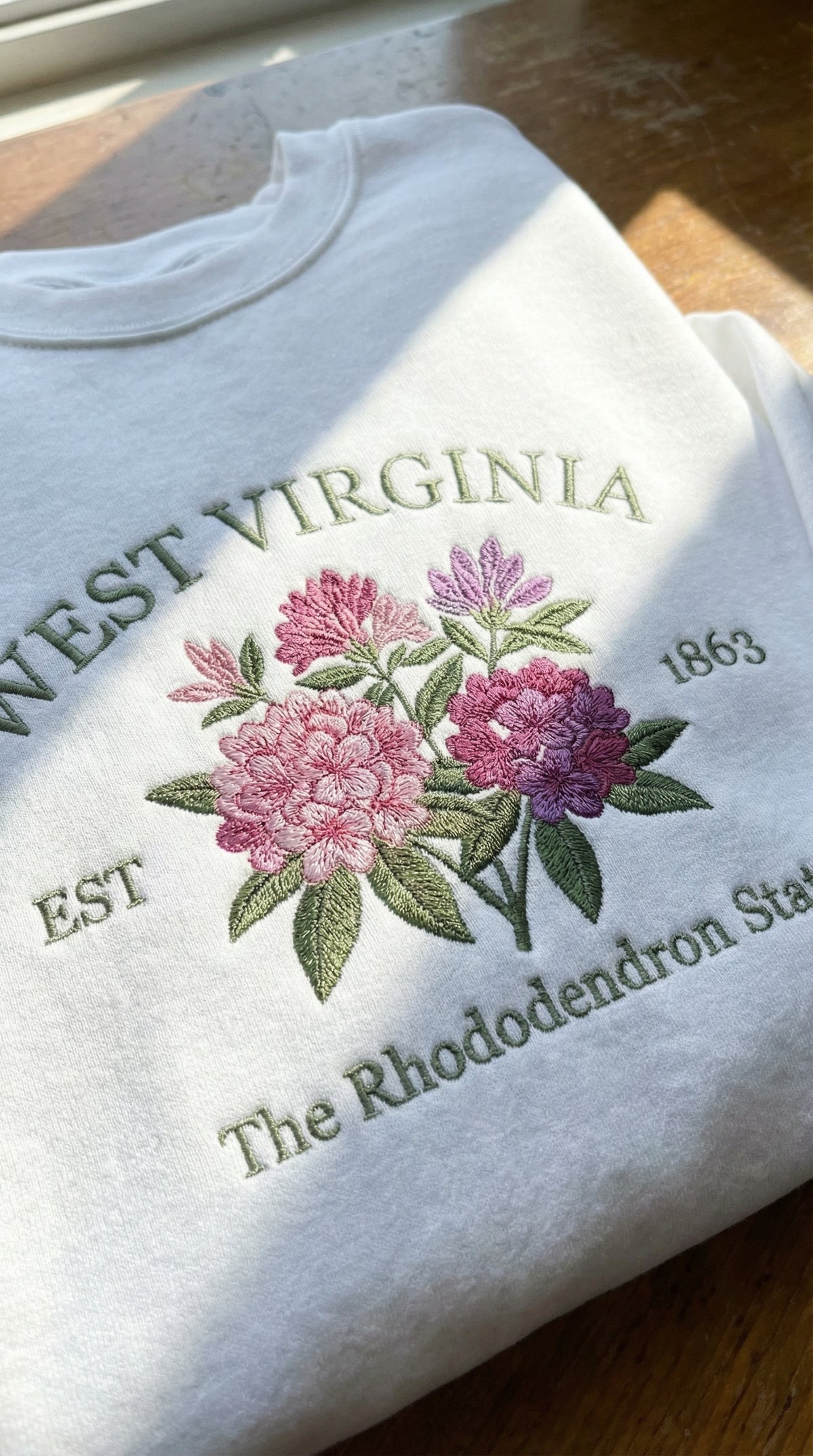White folded sweatshirt with detailed pink and purple Rhododendron flower embroidery, reading West Virginia EST 1863 The Rhododendron State