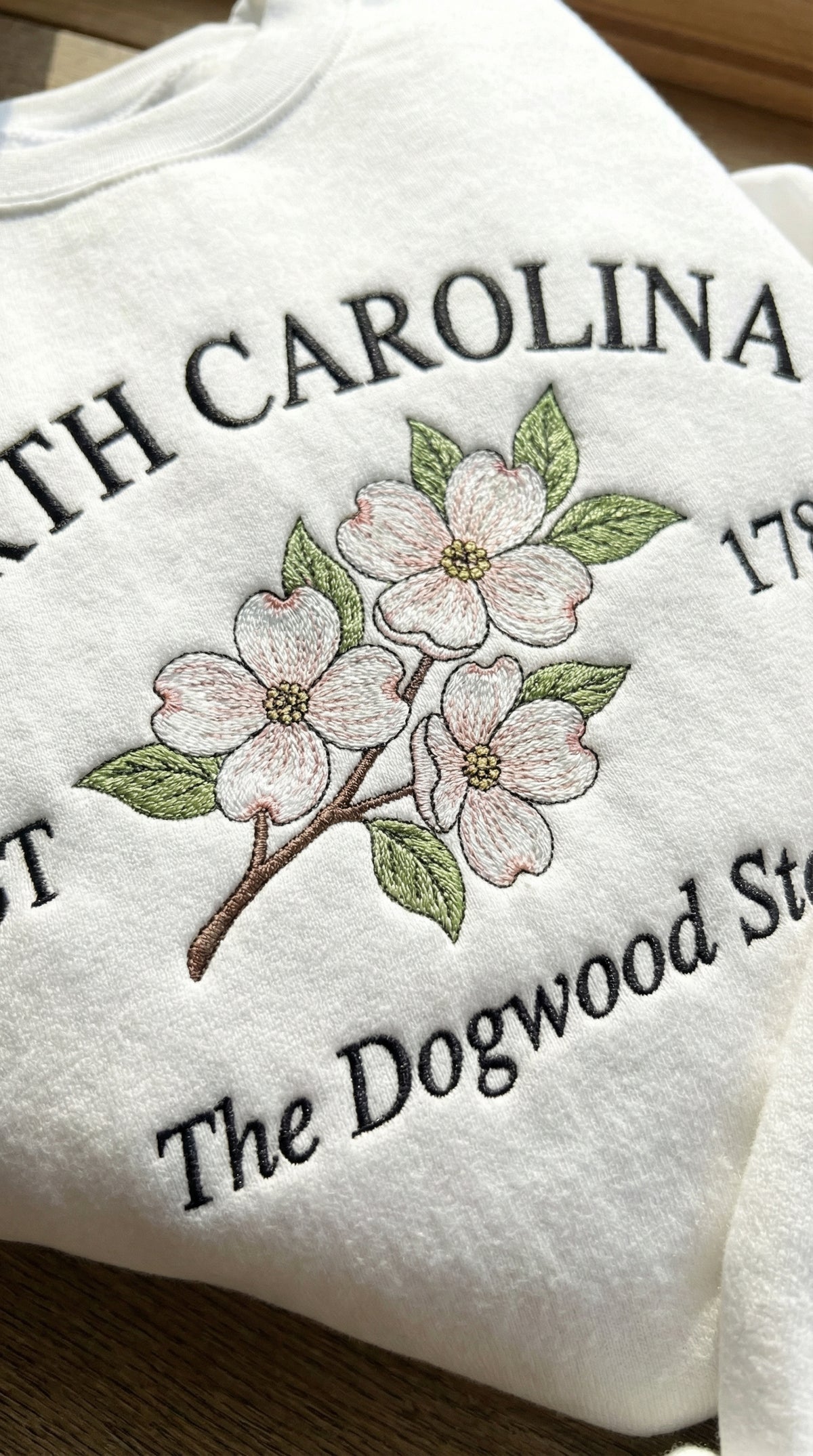 White folded sweatshirt with detailed pink and white Dogwood flower embroidery reading North Carolina EST 1789 The Dogwood State