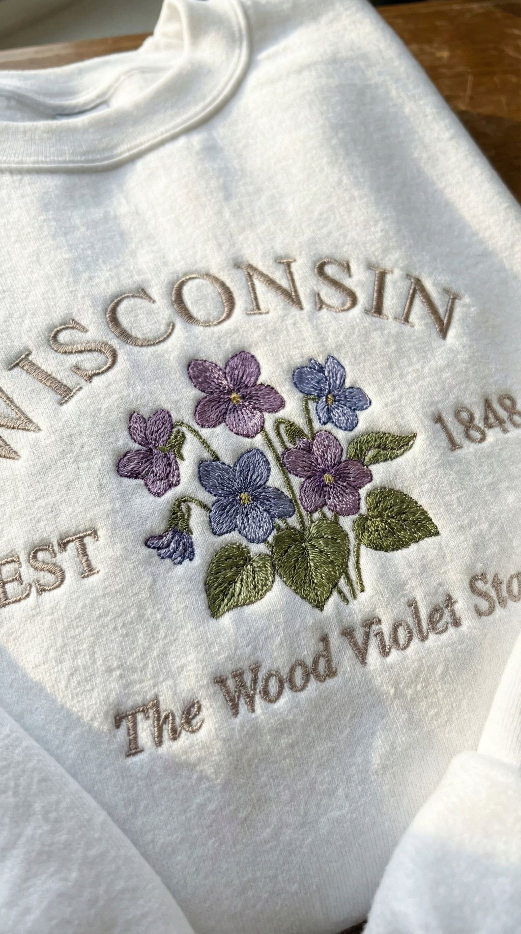 White folded sweatshirt with detailed purple Wood Violet flower embroidery reading Wisconsin EST 1848 The Wood Violet State