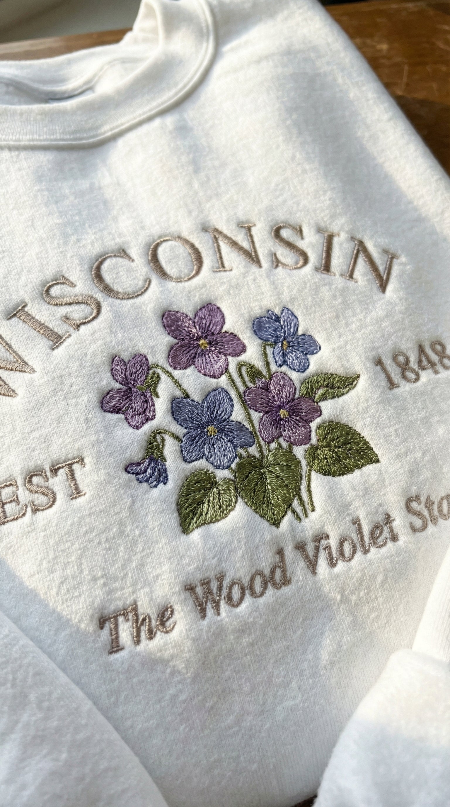 White folded sweatshirt with detailed purple Wood Violet flower embroidery reading Wisconsin EST 1848 The Wood Violet State