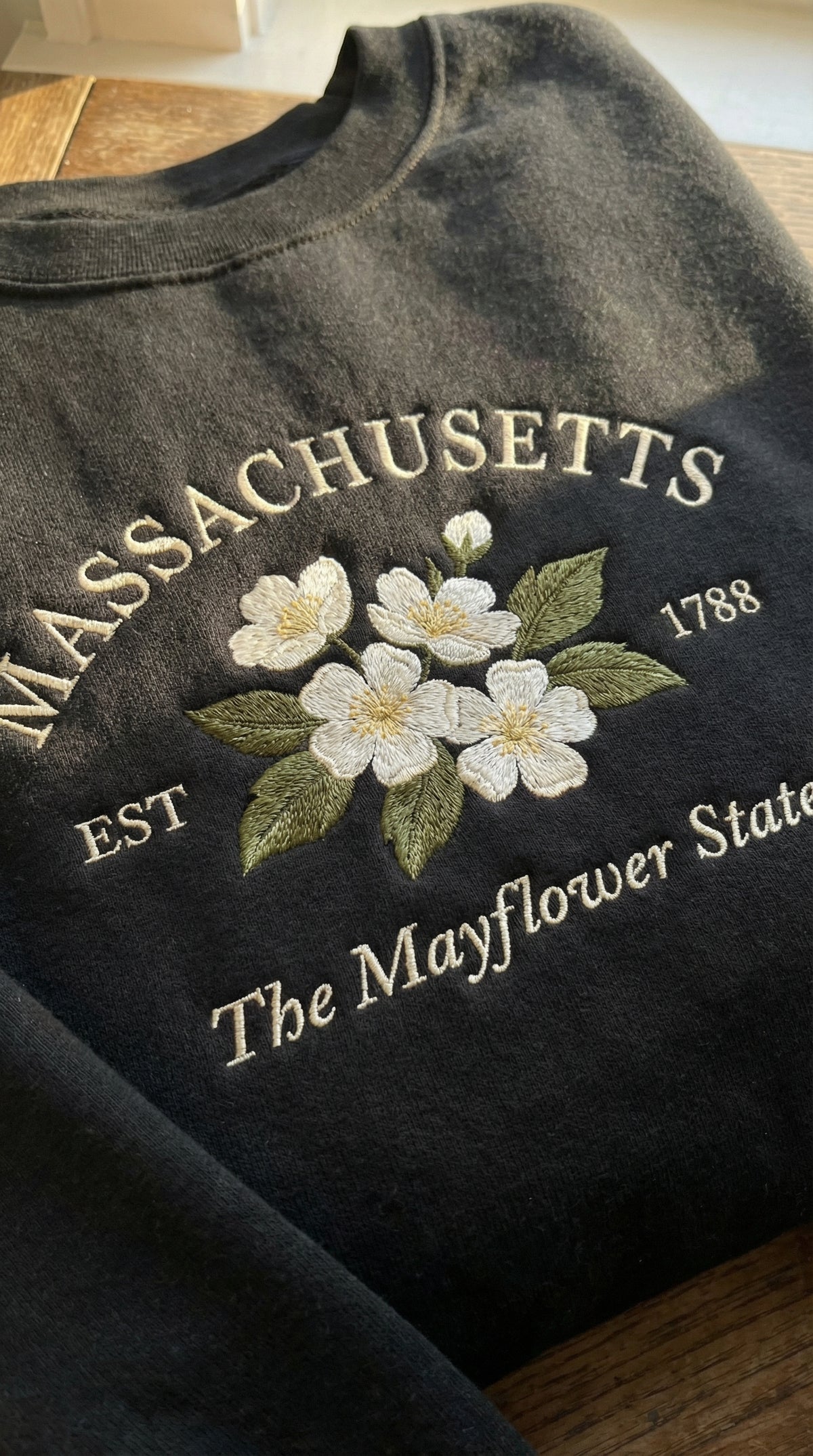 Black folded sweatshirt with detailed white Mayflower flower embroidery reading Massachusetts EST 1788 The Mayflower State