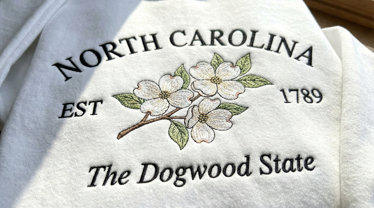 White folded sweatshirt with detailed pink and white Dogwood flower embroidery reading North Carolina EST 1789 The Dogwood State