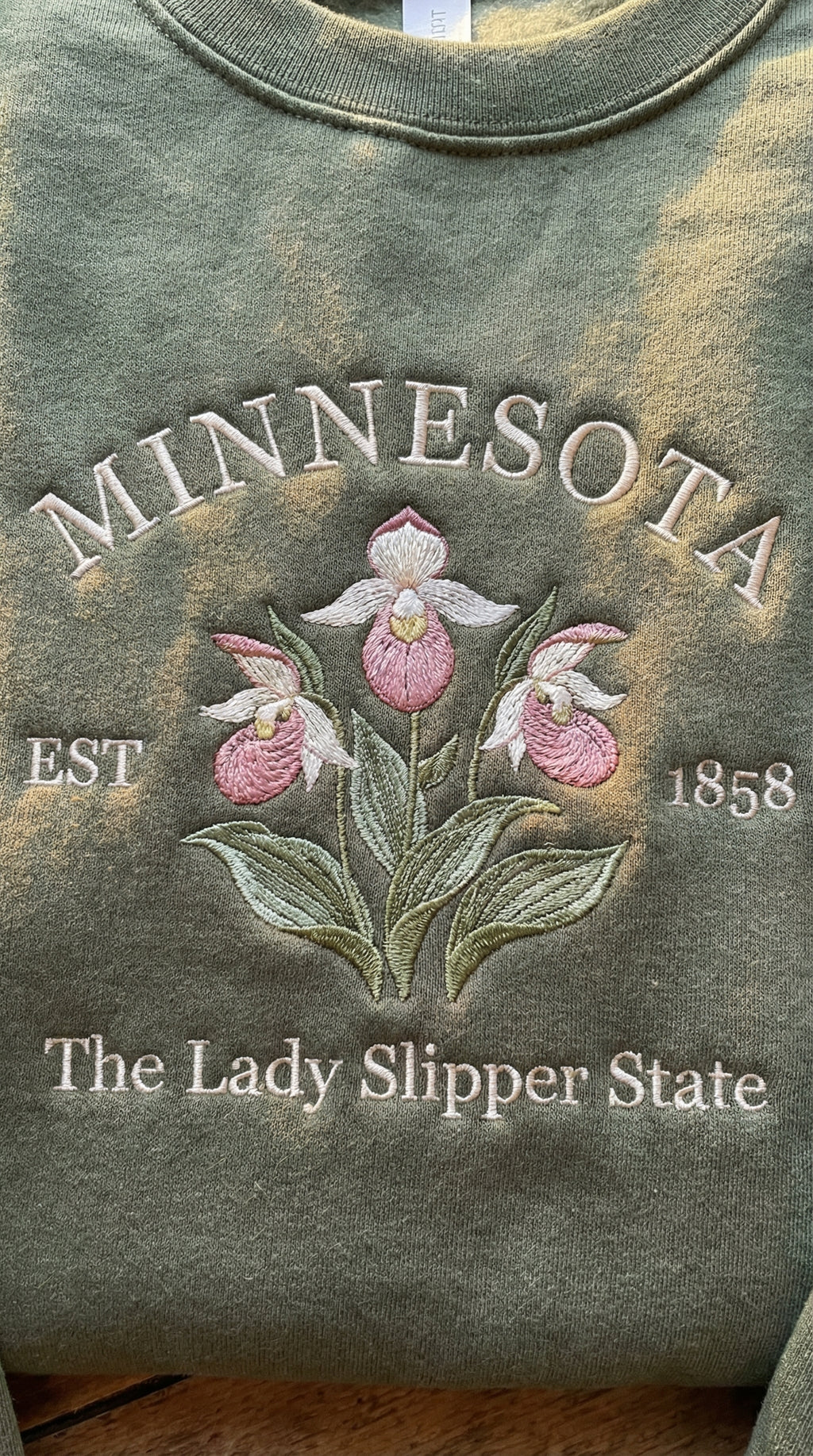 Olive green folded sweatshirt with detailed pink and white Lady Slipper orchid embroidery reading Minnesota EST 1858 The Lady Slipper State