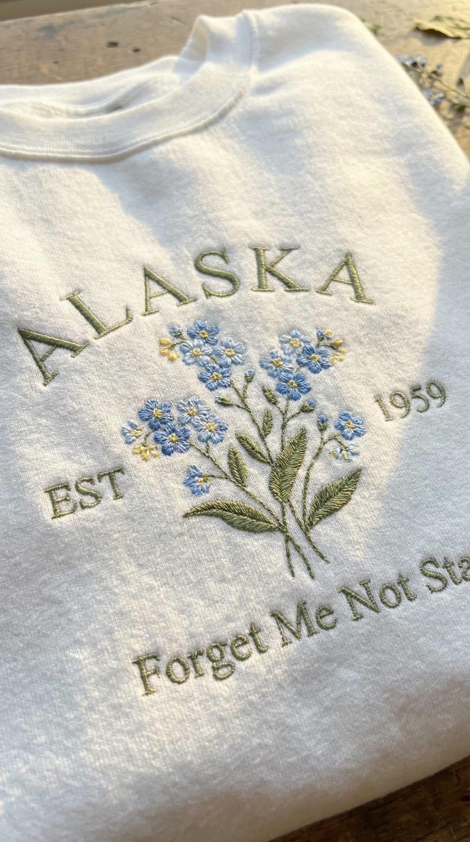 White folded sweatshirt with detailed blue Forget-Me-Not flower embroidery reading Alaska EST 1959 Forget Me Not State