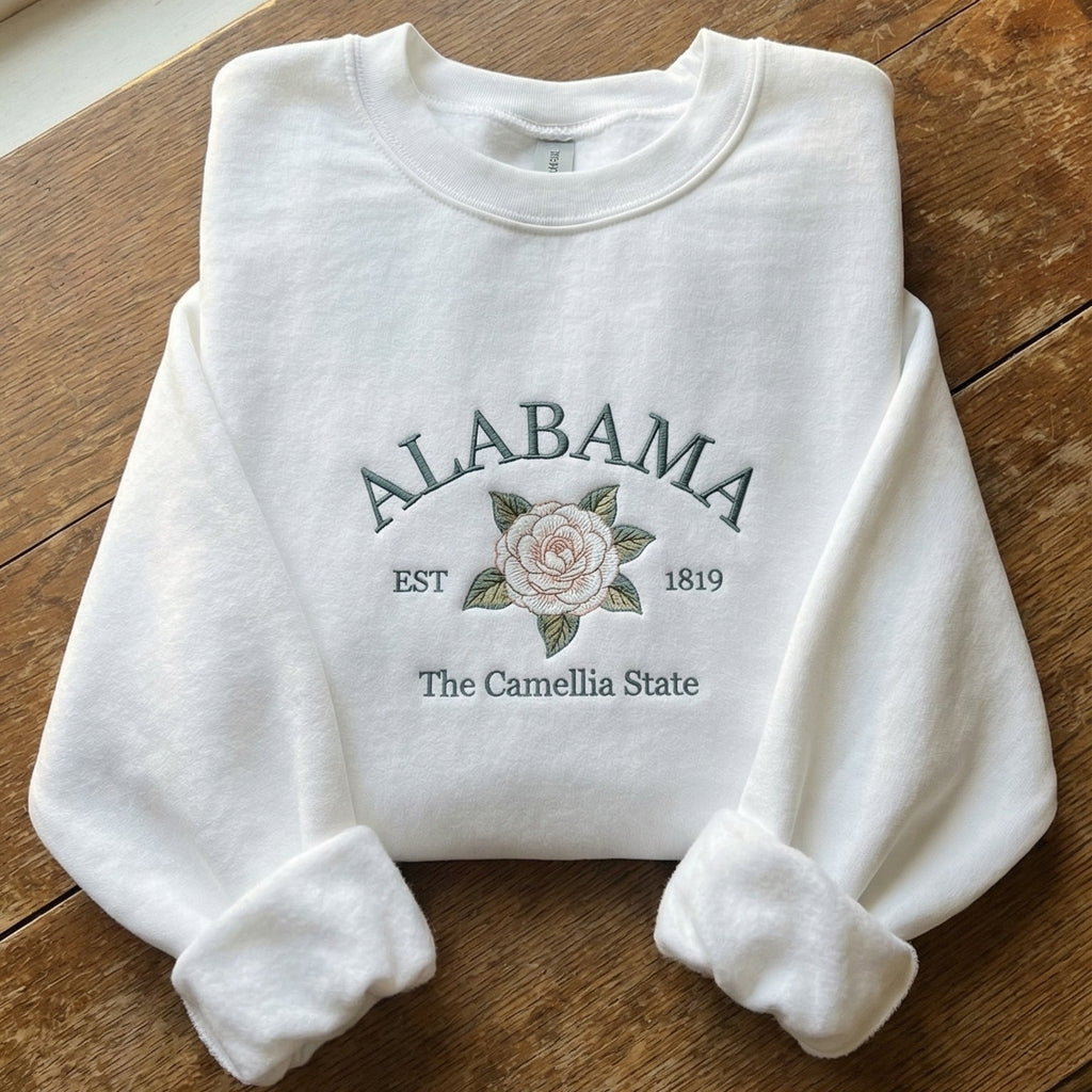 White folded sweatshirt with detailed pink Camellia flower embroidery and sage green text reading Alabama EST 1819 The Camellia State