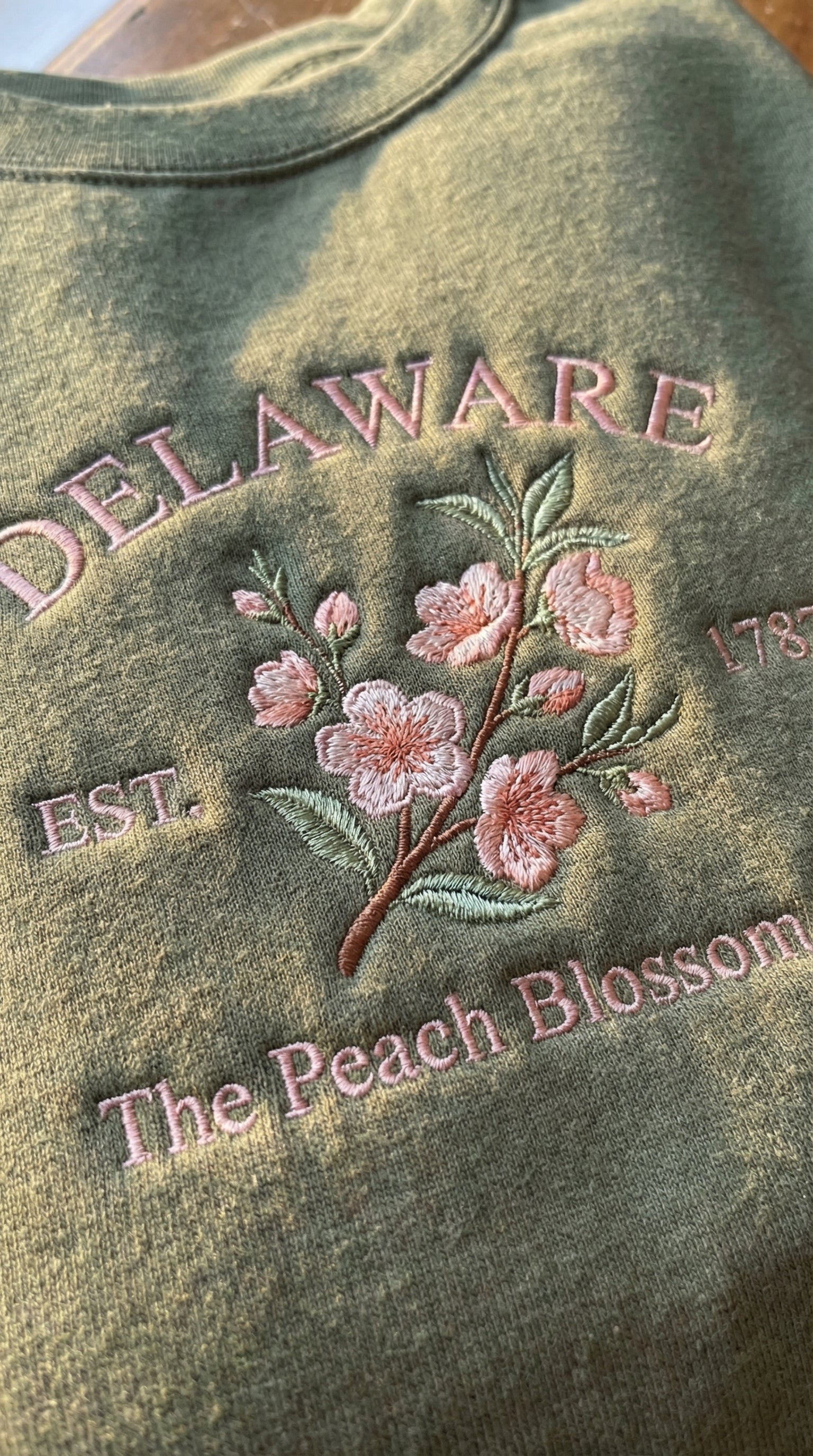 Olive green sweatshirt with detailed pink Peach Blossom flower embroidery reading Delaware EST. 1787 The Peach Blossom State